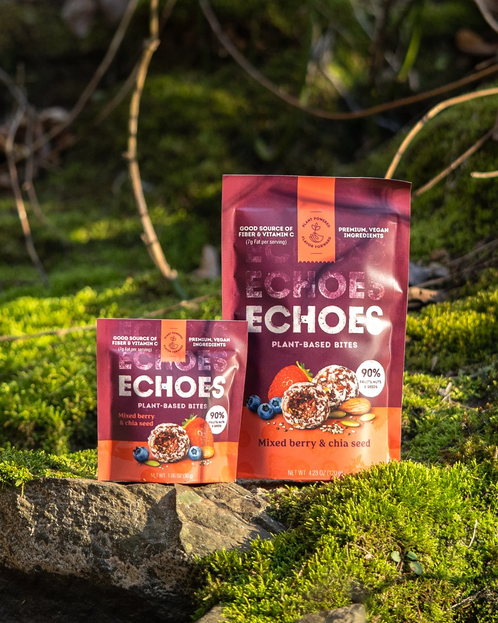 Two bags of Echoes plant-based bites, with one smaller and one larger, placed on mossy ground outdoors. The packaging is pink and orange, featuring images of mixed berries and chia seeds.