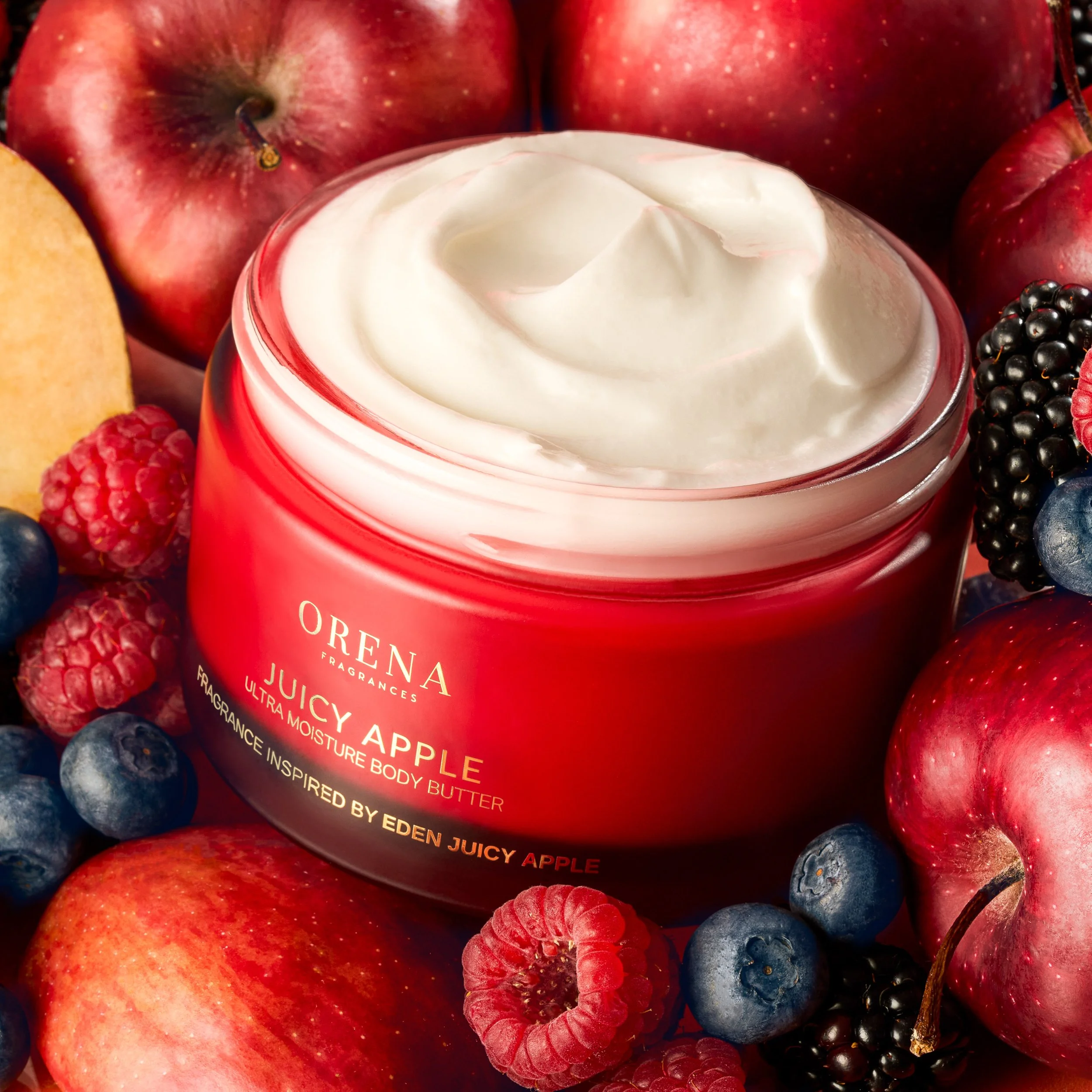 A red jar of ORENA Juicy Apple body butter surrounded by apples, raspberries, blueberries, blackberries, and an apple slice.