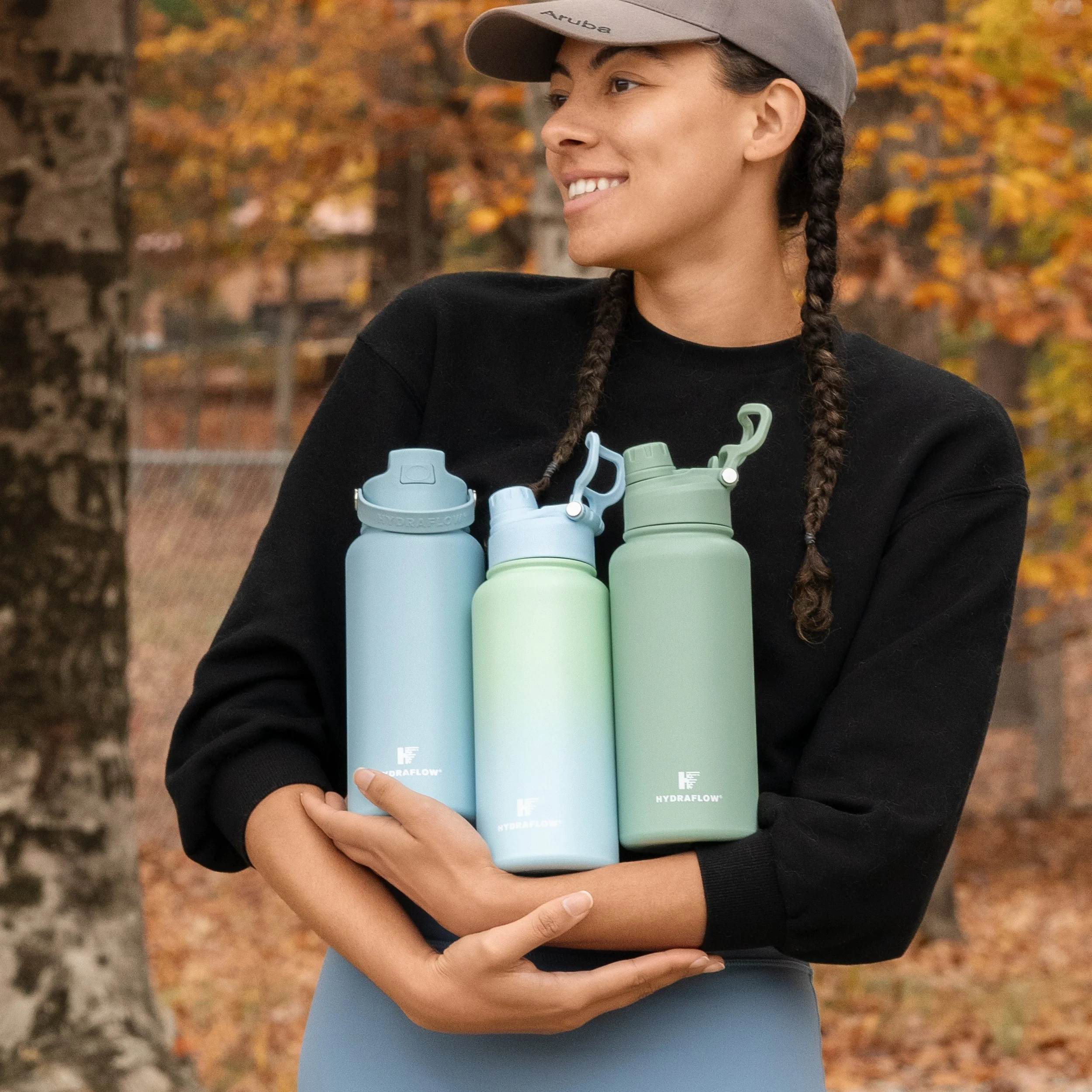 A woman holding three pastel-colored water bottles against a backdrop of autumn trees with fall foliage.