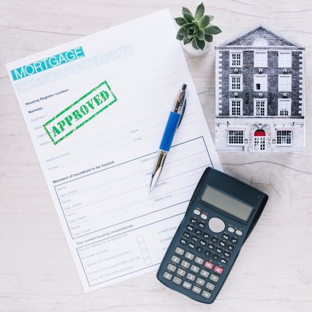 A flat lay image showing a mortgage application form with a green 'APPROVED' stamp, a blue pen, a small calculator, and a miniature model of a dark-colored townhome or apartment building.