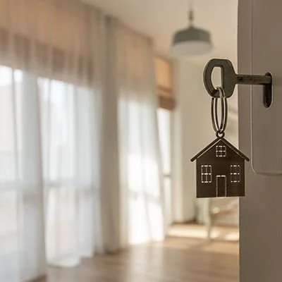 A close-up shot of a house-shaped keychain attached to a key in a lock, opening a door to a bright, empty room with hardwood floors and sunlit sheer curtains.