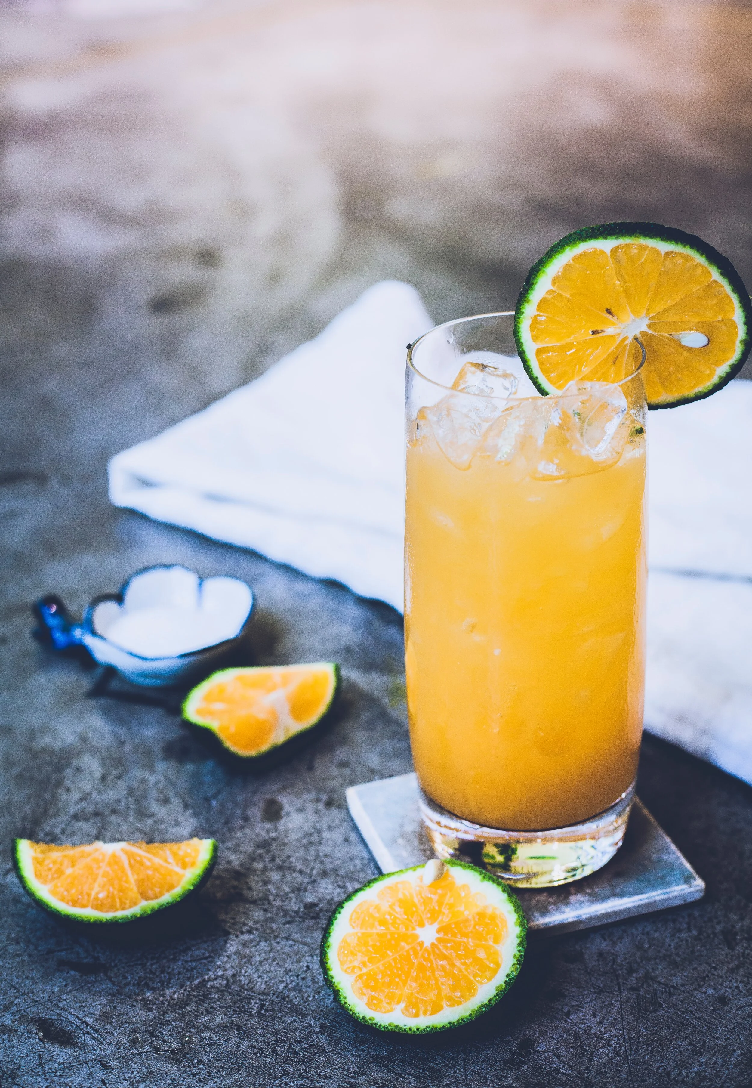 A tall glass of orange beverage with ice, garnished with a slice of lime and orange, on a coaster, with extra lime and orange slices and a white napkin in the background.