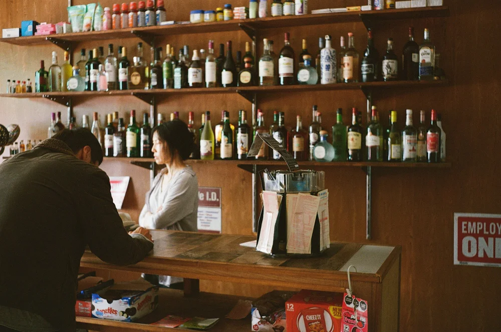 Still from Scratcher: a man at a corner-store counter, the cashier behind it