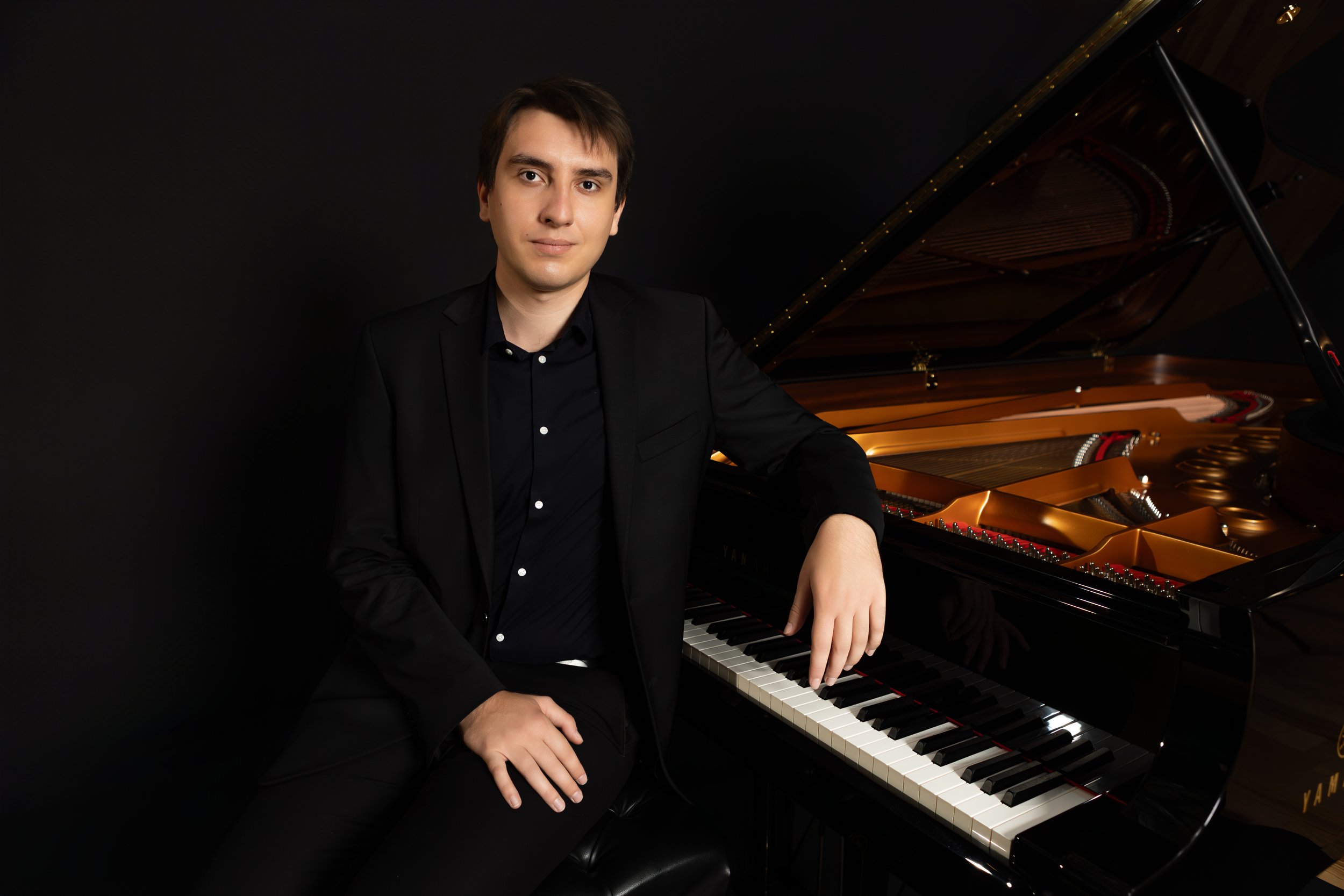 A young man in a black suit sitting at a grand piano, looking at the camera with a neutral expression.