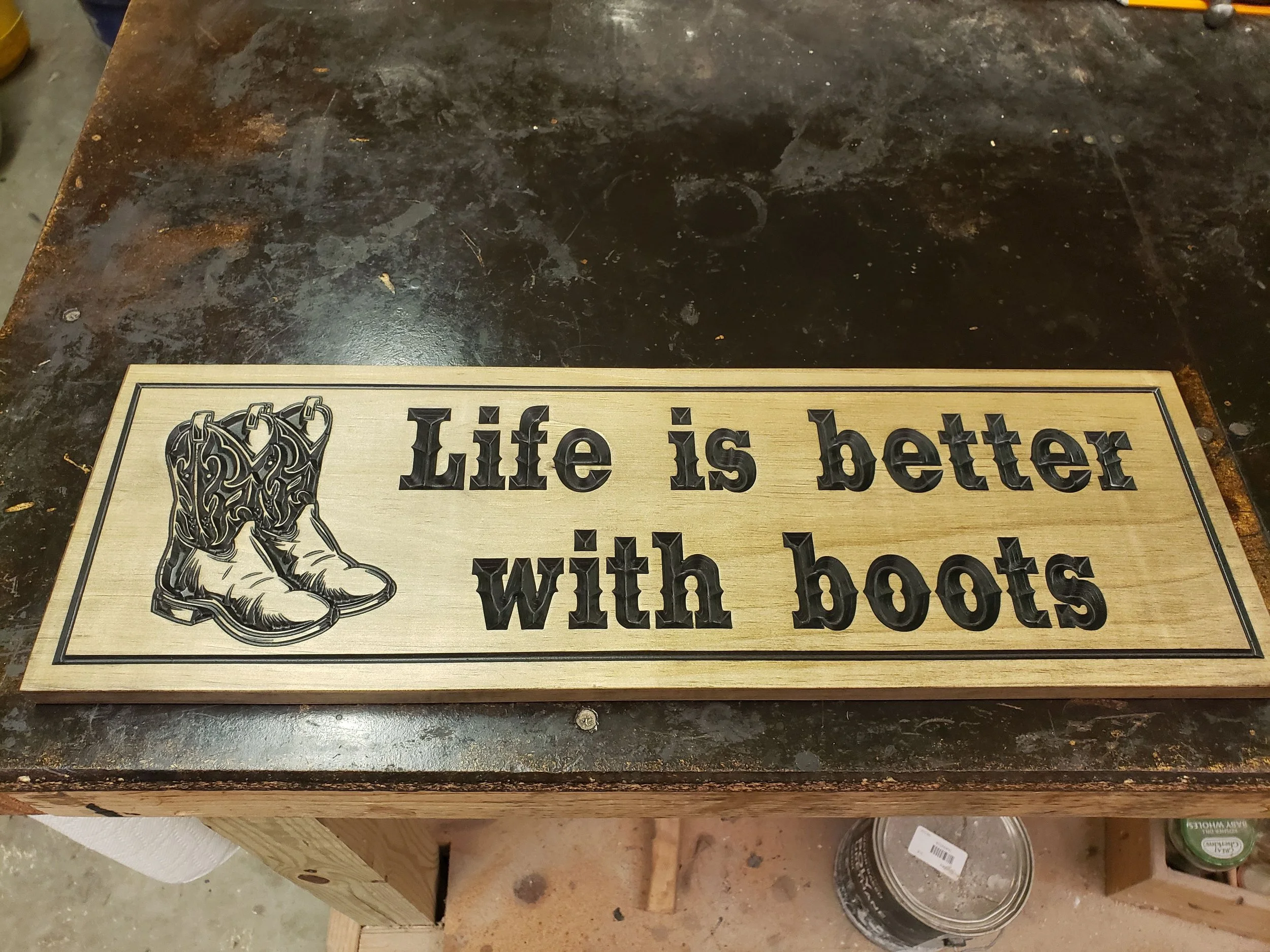Rectangular wooden wall sign featuring detailed carved cowboy boots on the left and the phrase 'Life is better with boots' in bold, raised black lettering on the right, resting on a dark workbench.