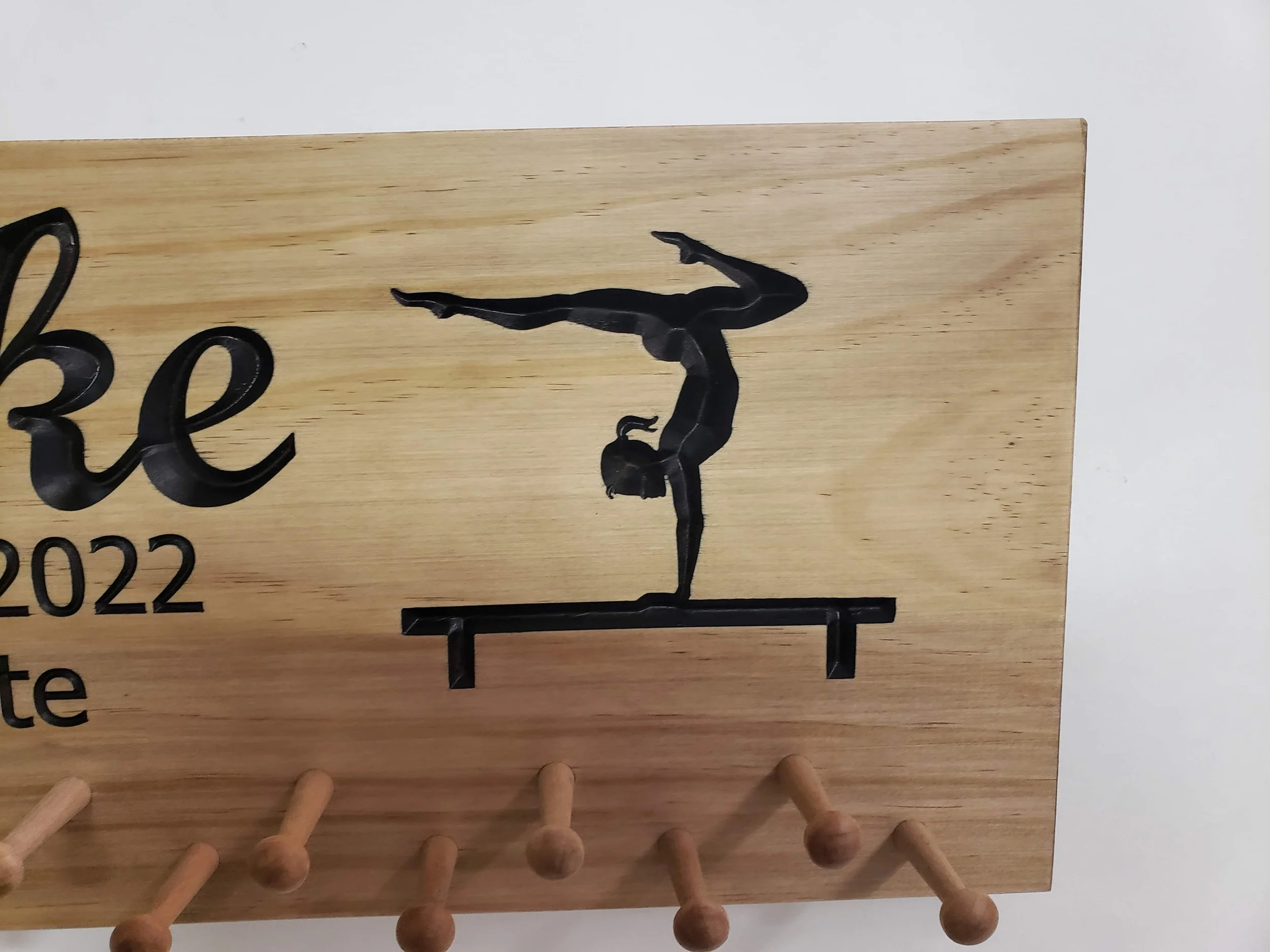 Close-up of the right side of a custom gymnastics medal holder showing a detailed black gymnast silhouette performing on the balance beam, with part of the personalized text visible. Natural wood finish with sturdy wooden pegs below.