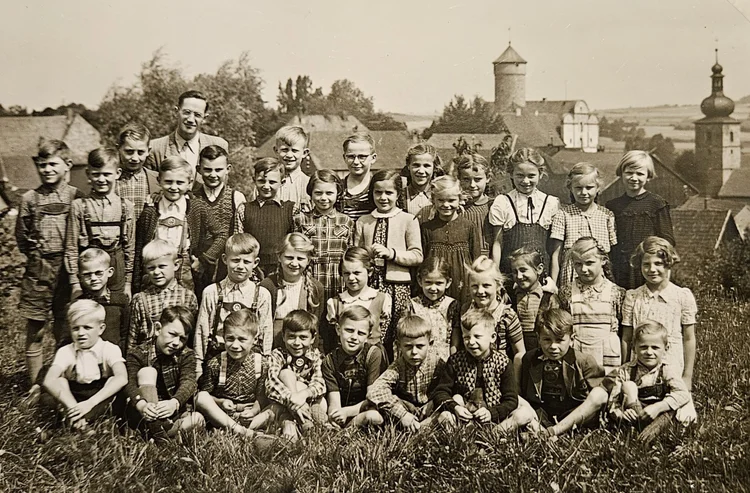 Black and white photograph of a group of school children and their teacher outdoors, with houses and church steeples in the background.