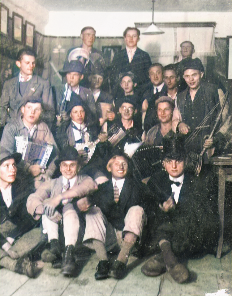 A black-and-white vintage photo of a group of people, some holding musical instruments, in a room with framed pictures on the wall.