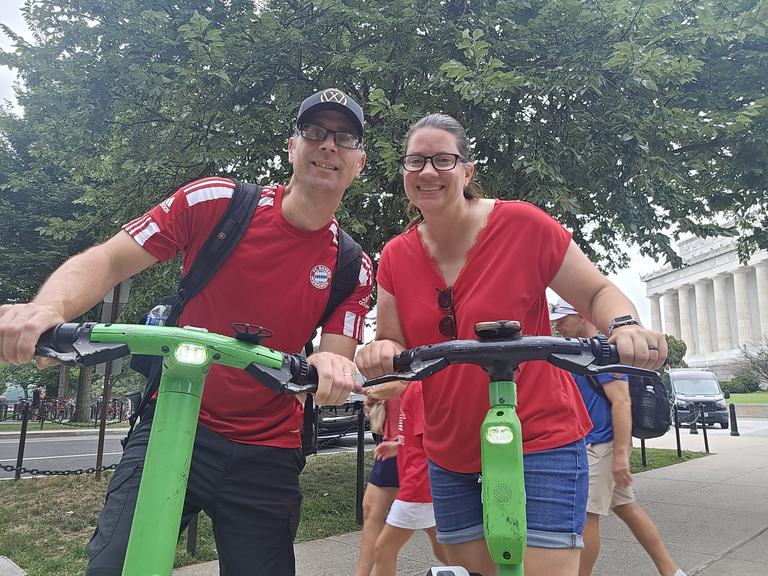 Two people, a man and a woman, smiling and holding onto bright green electric scooters outdoors on a cloudy day. They are wearing red shirts and glasses, with trees and a historic building in the background.