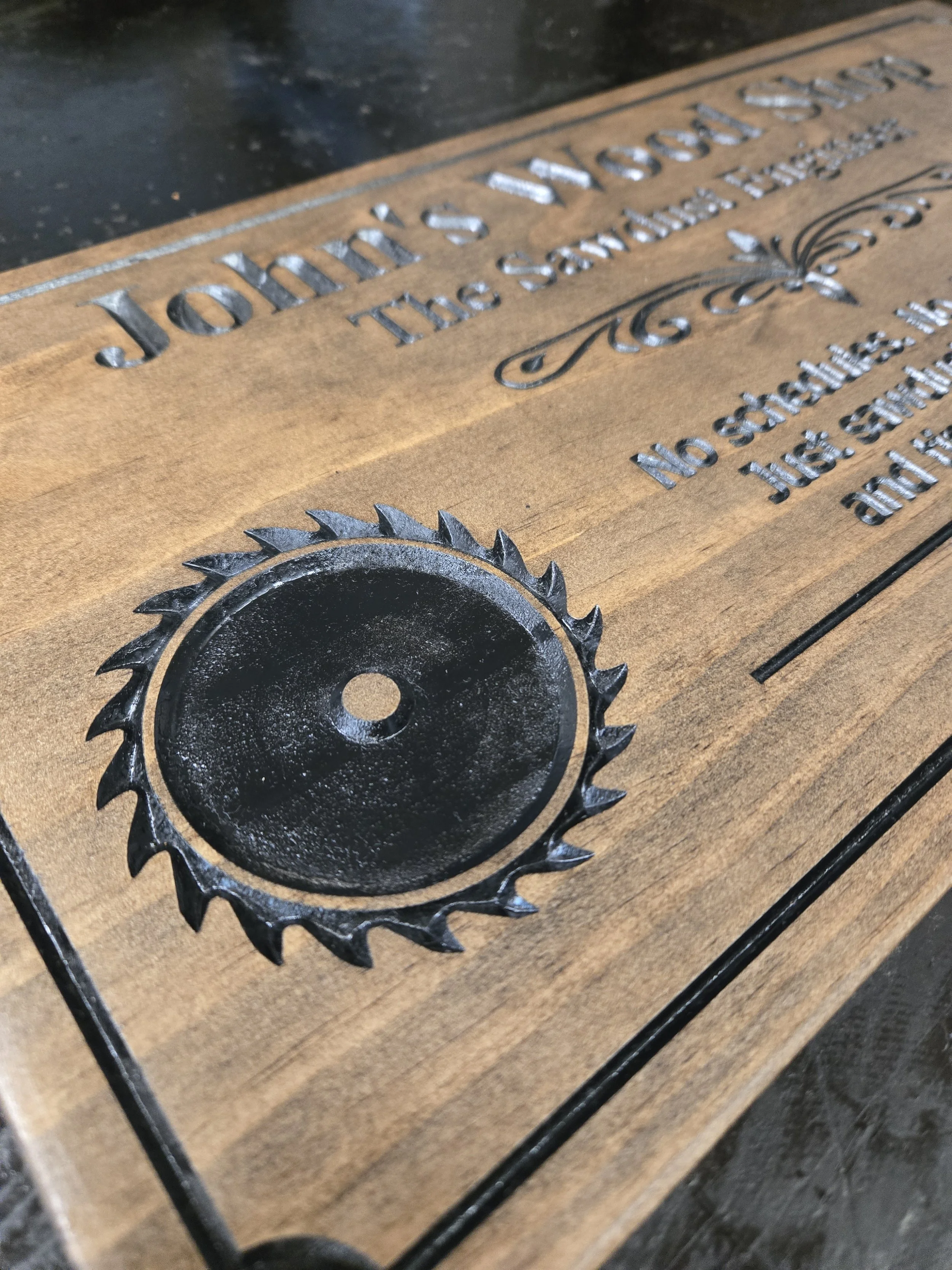 Close-up of a wooden plaque featuring a large engraved circular saw blade on the left, with “John’s Wood Shop” and “The Sawdust Engineer” above, plus partial text “No schedules. No deadlines. Just sawdust…” on the right
