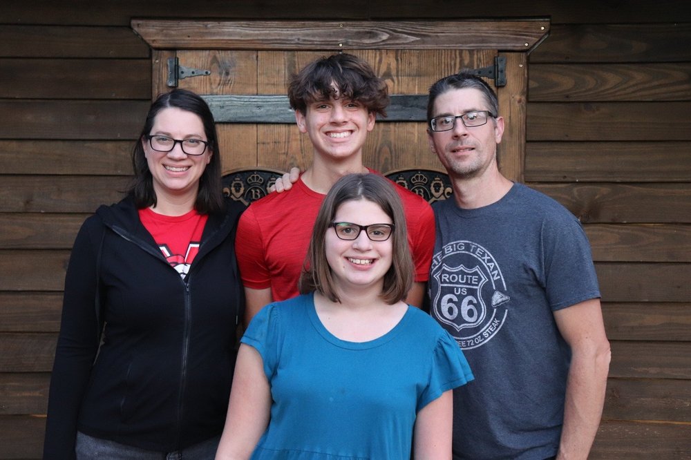 Family portrait of four people standing outdoors in front of a wooden wall, smiling at the camera.