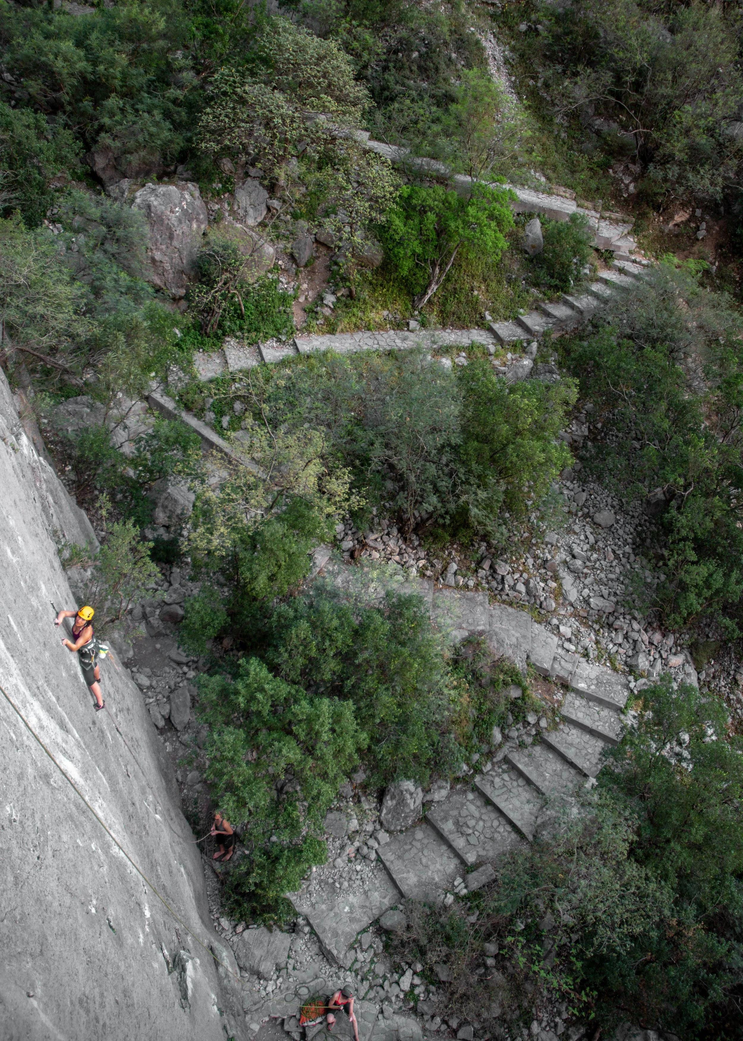 Rock climbers on a vertical cliff near a winding stone staircase surrounded by greenery and rocky terrain.