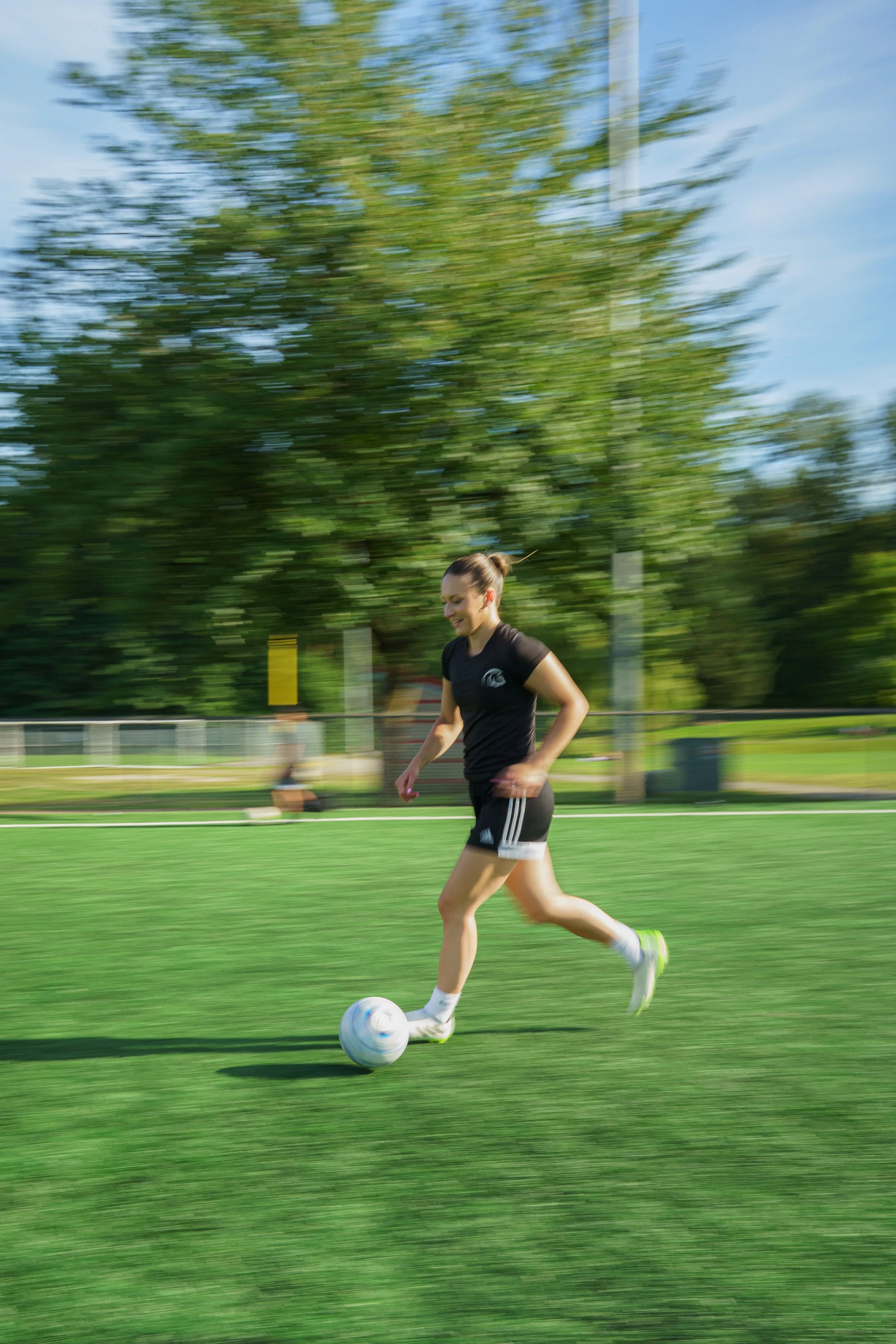 A young woman playing soccer on a grassy field, with trees and a blue sky in the background.