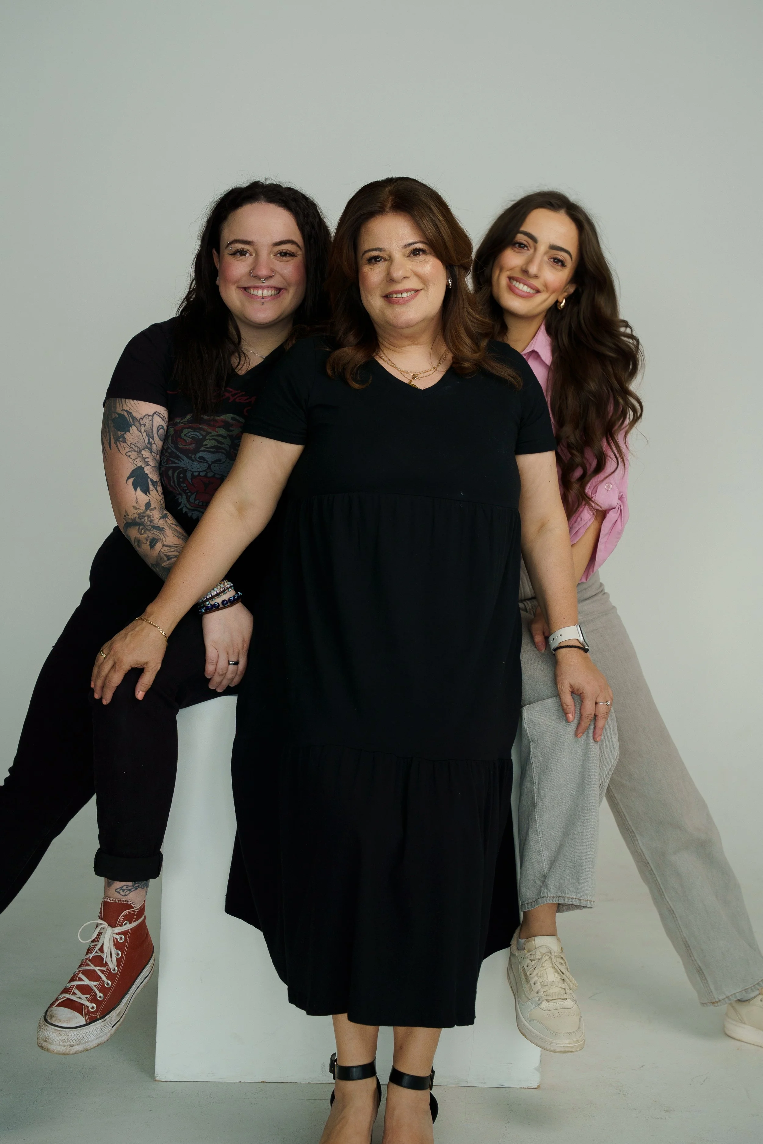 Three women smiling together against a plain background. One woman is seated on a white block, and the other two are standing beside her. They are dressed casually.