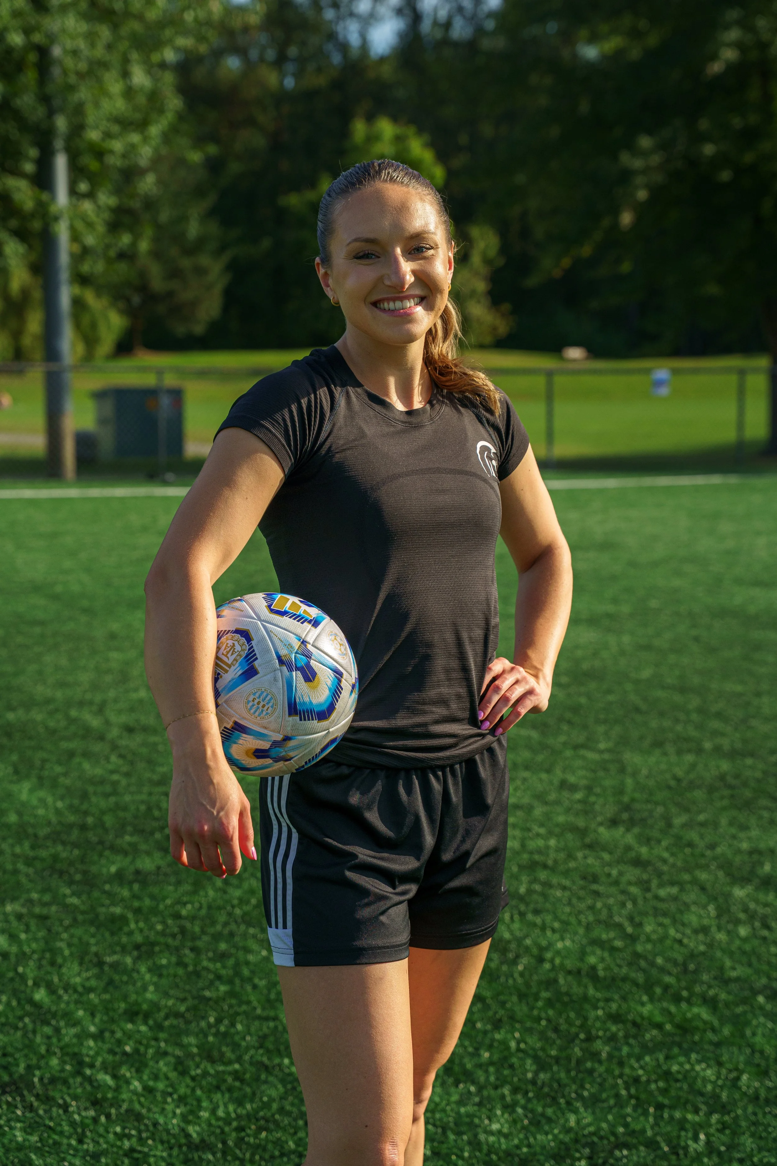 A smiling woman in athletic clothing holding a soccer ball on a grassy field.