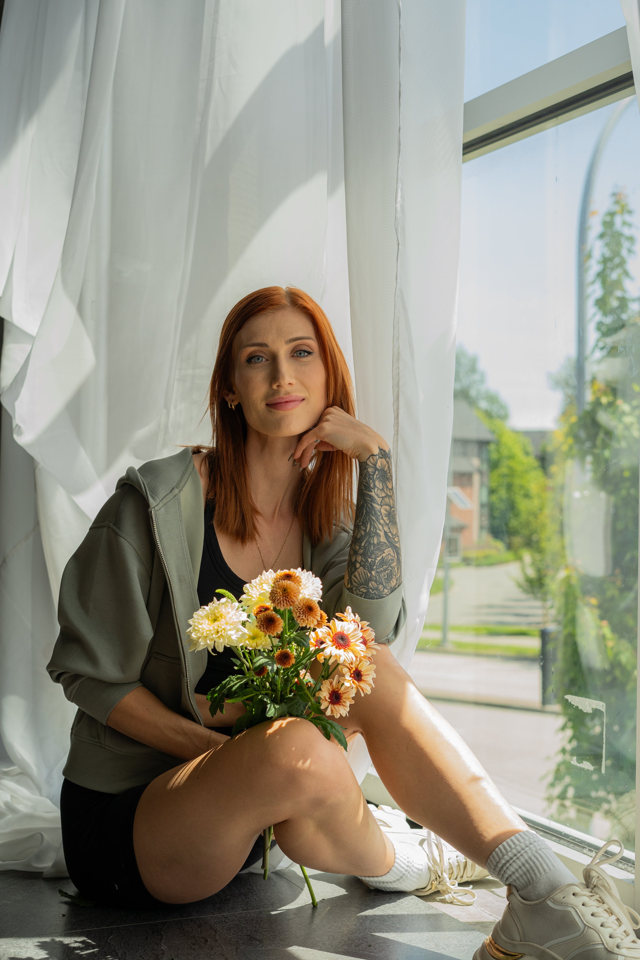 Red-haired woman sitting by a large window with sheer white curtains, holding a bouquet of flowers, wearing a black shirt, gray hoodie, shorts, and sneakers, with sunlight illuminating her face and surroundings.