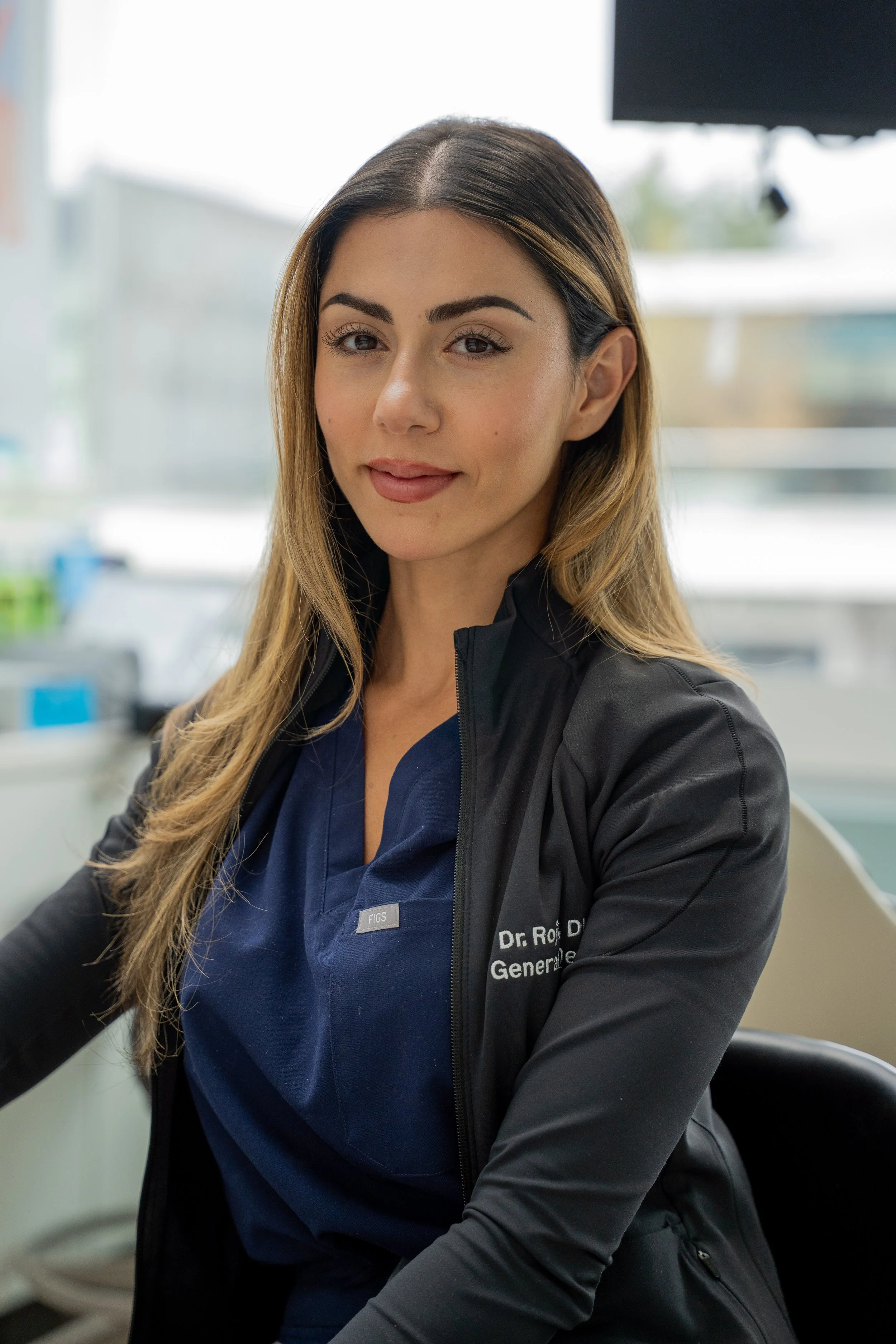 A woman at a veterinary clinic with long brown hair, wearing a blue scrubs and a black jacket with her name and title embroidered, sitting in front of a window with a blurred background of medical equipment.