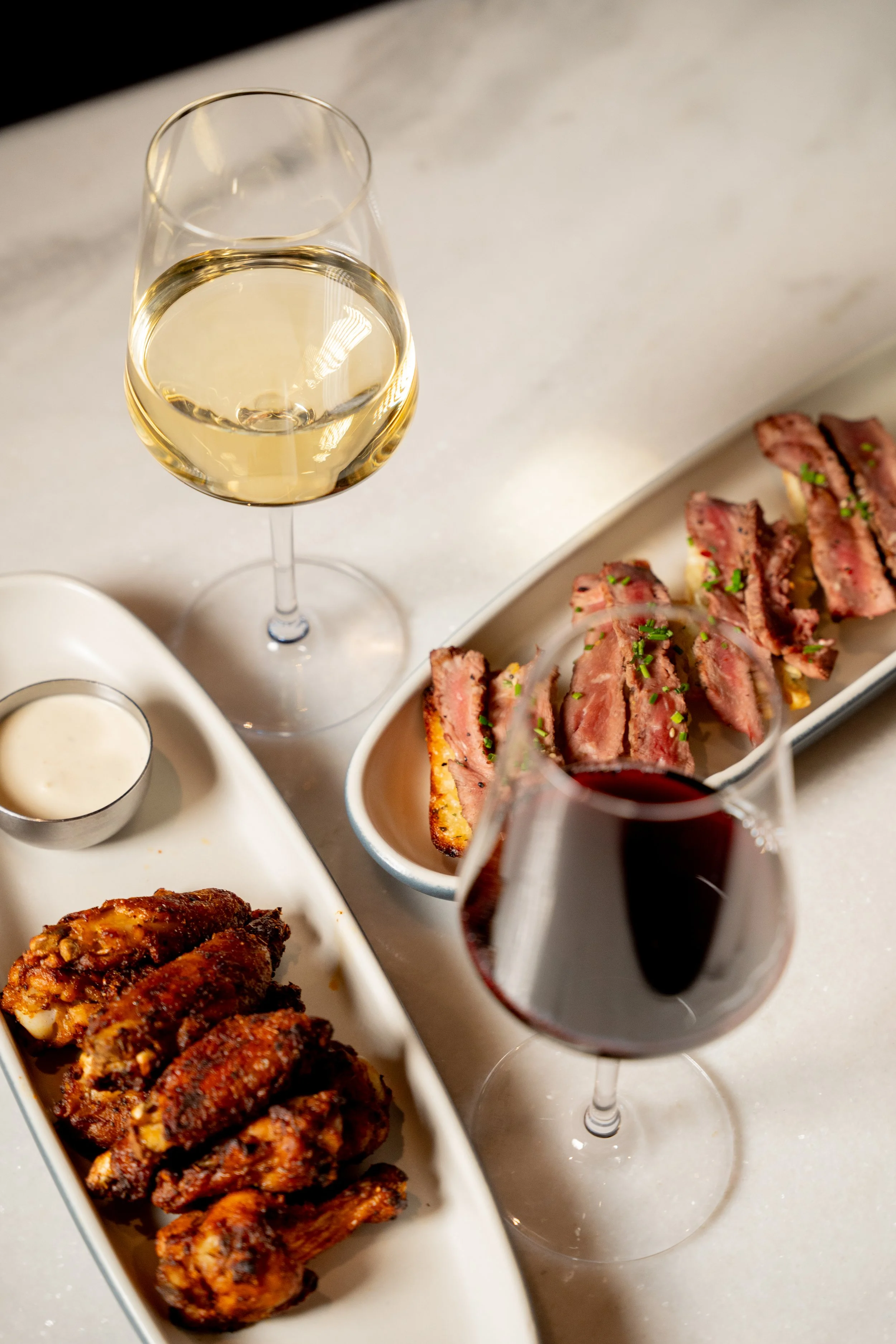 A plate of grilled chicken wings, a glass of white wine, a glass of red wine, and a plate of sliced steak with green herbs on top.