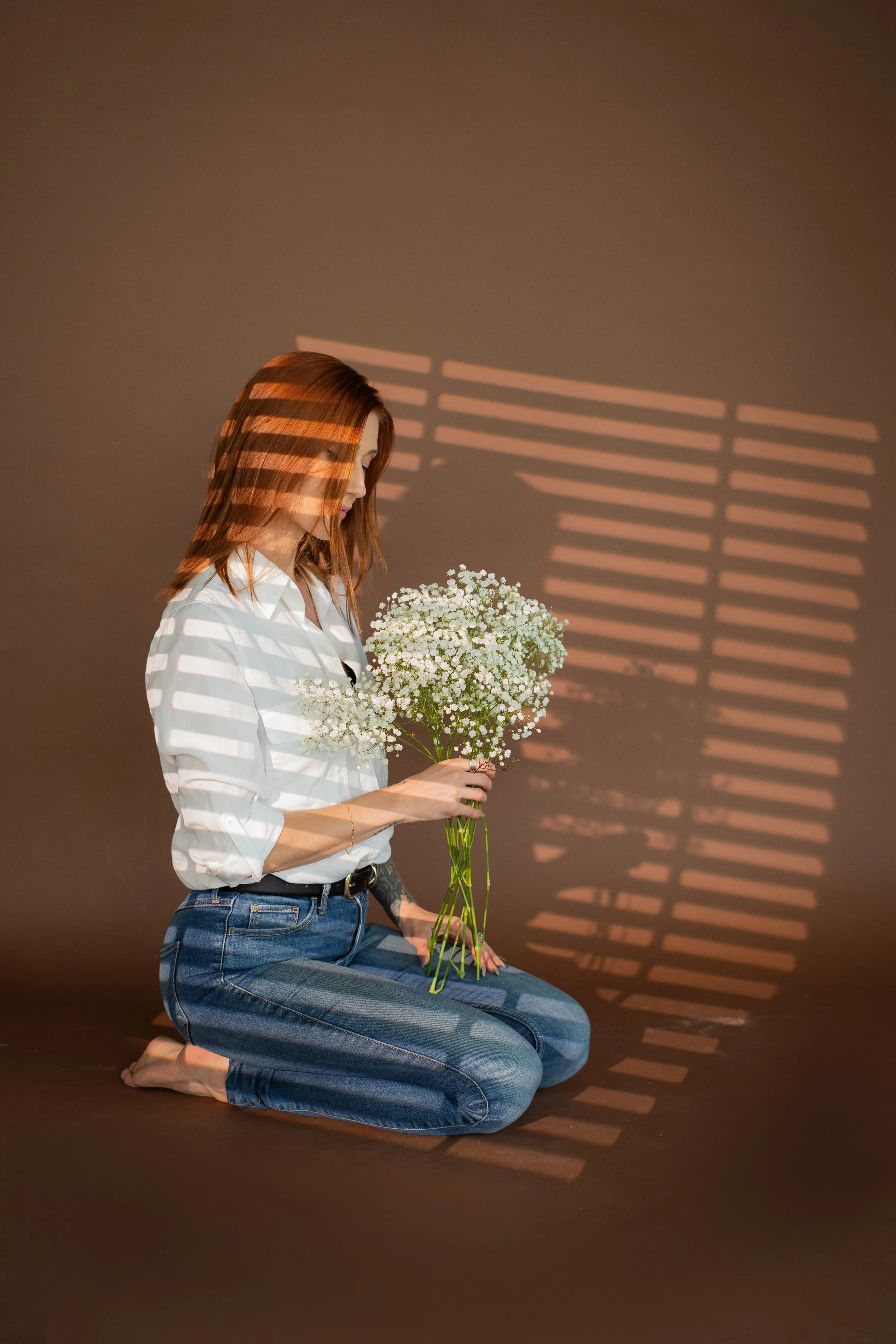 A woman with red hair, wearing a white shirt and jeans, kneeling on the floor, holding a bouquet of white flowers. Shadows from window blinds create striped patterns on the brown wall behind her.