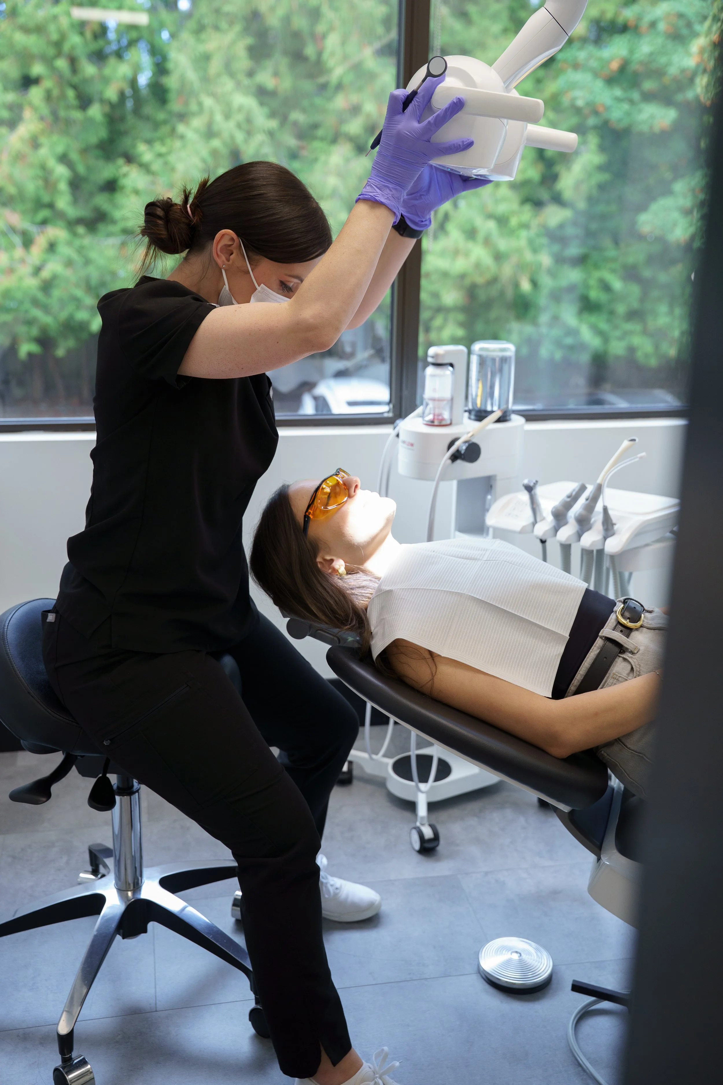 A dentist wearing purple gloves and a face mask is performing a dental procedure on a patient lying in a dental chair with sunglasses on, in a dental office with large windows and dental equipment.