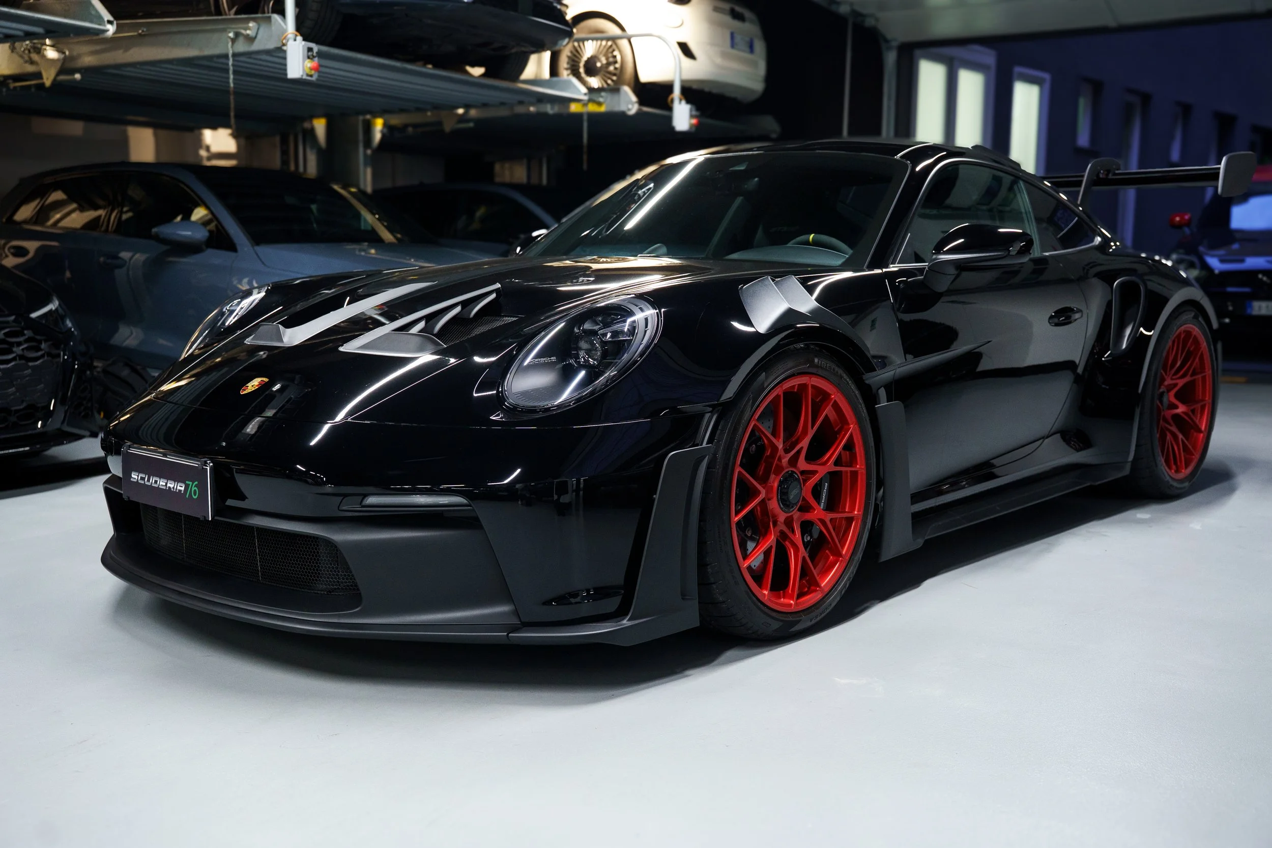 A black Porsche racing car with red rims parked in an indoor garage with other cars in the background.