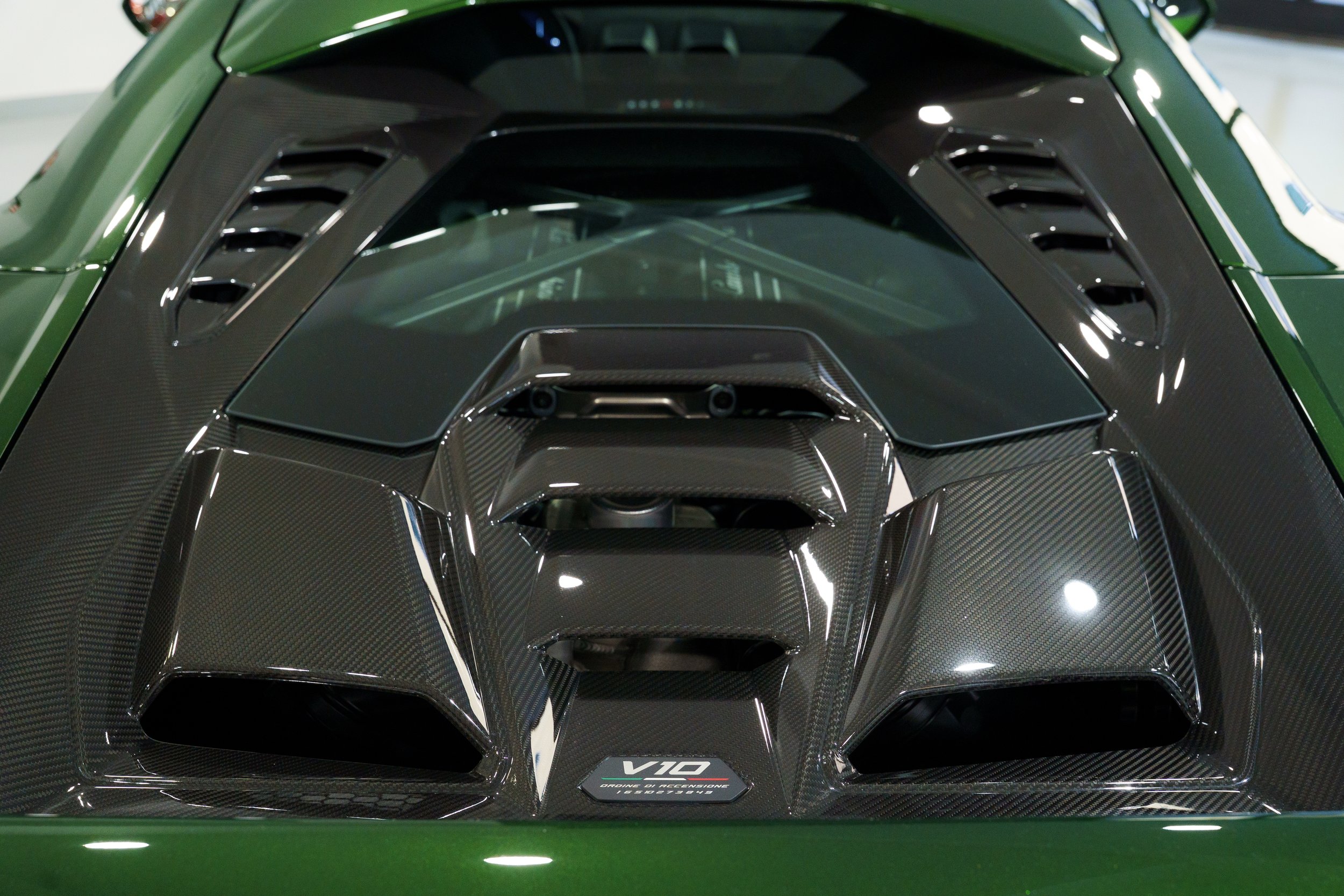 Close-up view of the rear engine cover of a green Lamborghini, showcasing a transparent window and carbon fiber engine cover with 'V10' badge.