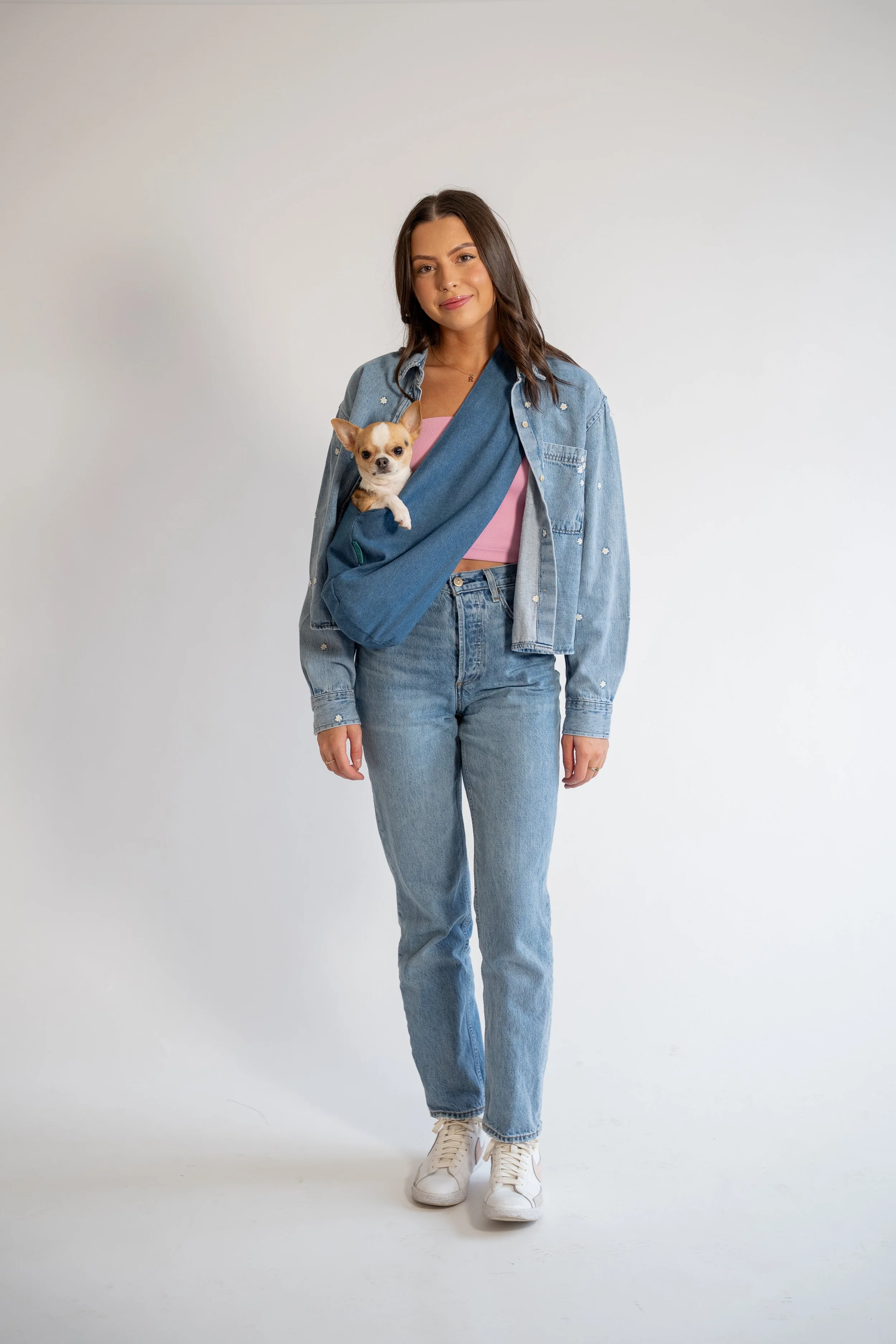A woman with long dark hair wearing a denim jacket and jeans, standing against a plain white background. She has a pink top underneath and is carrying a small dog in a blue sling across her chest.