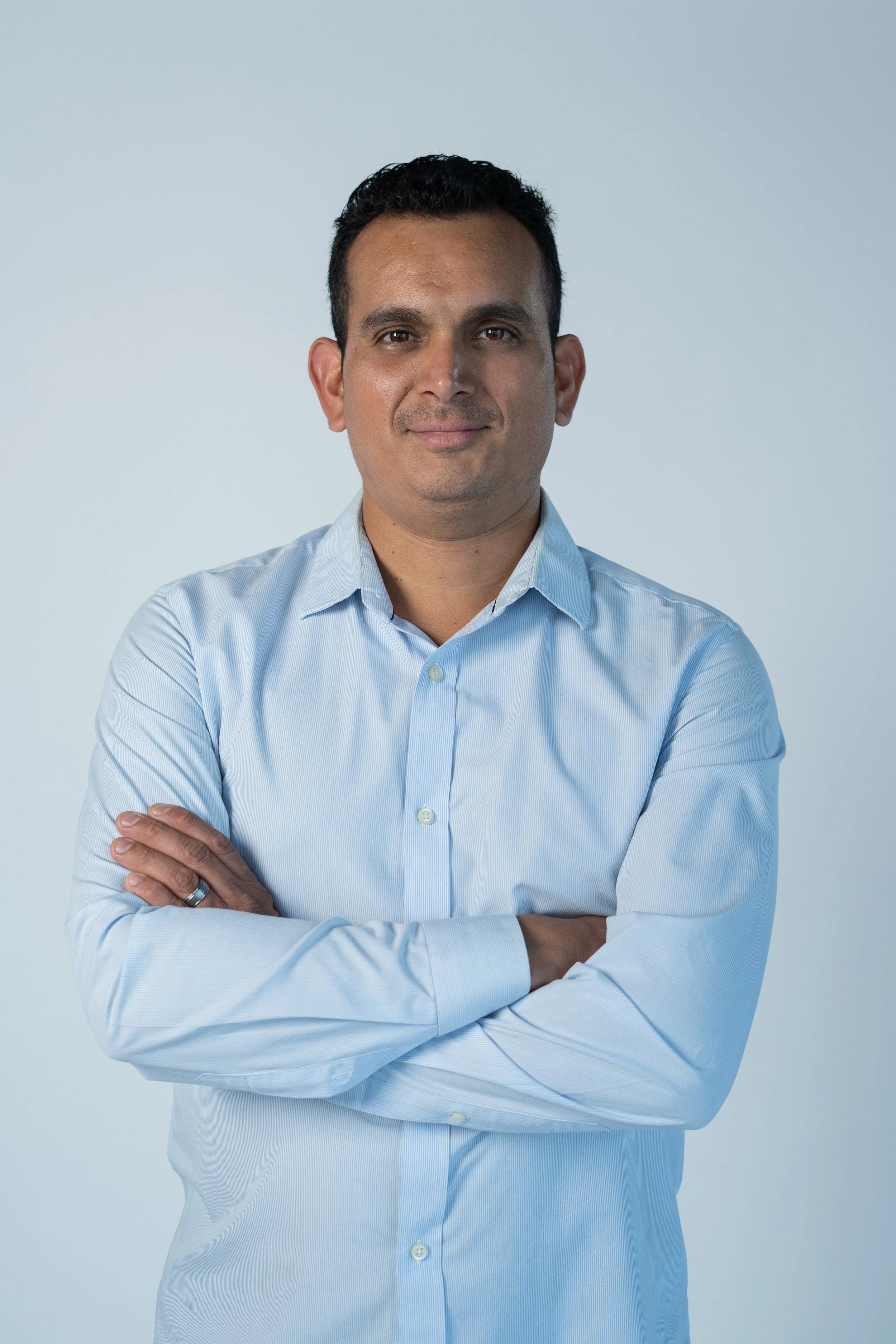 A man in a light blue button-up shirt with arms crossed, looking at the camera against a plain light background.