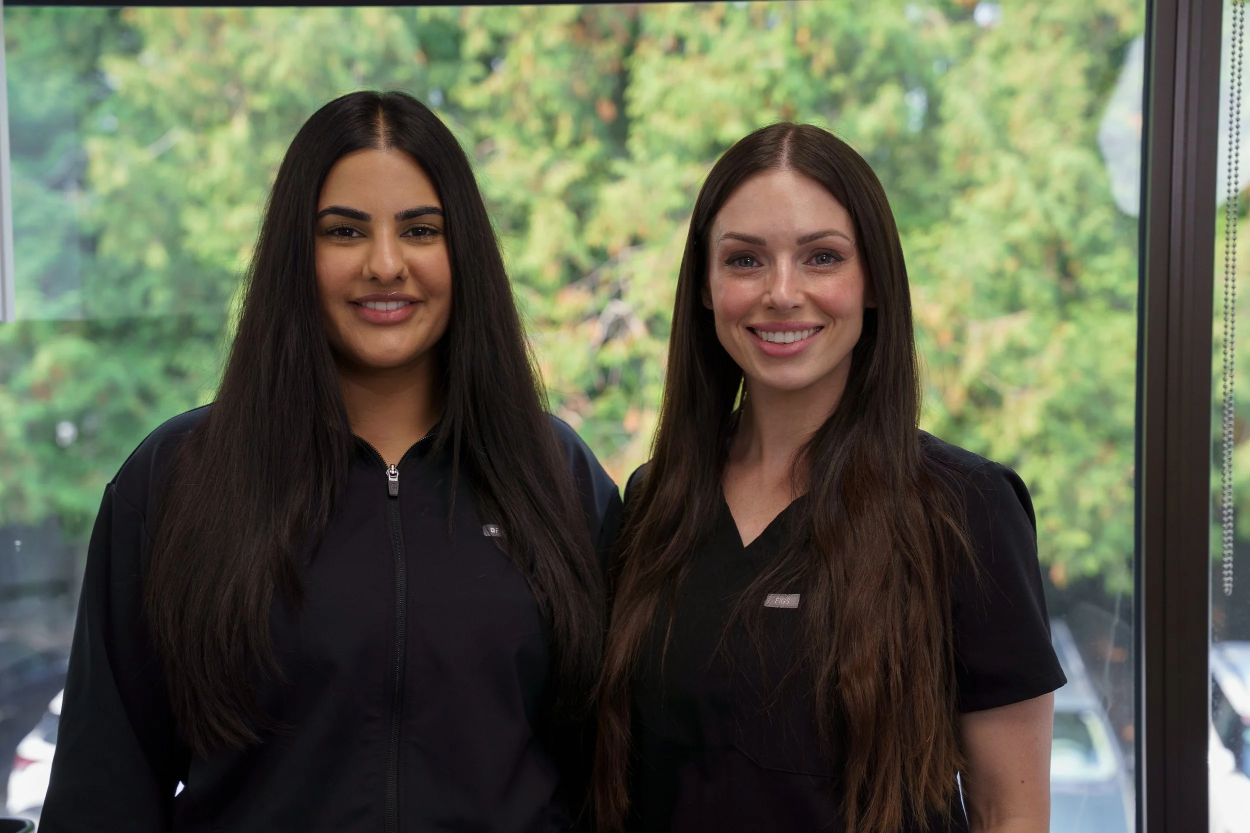 Two women standing indoors in front of a window with green trees outside, both smiling at the camera.