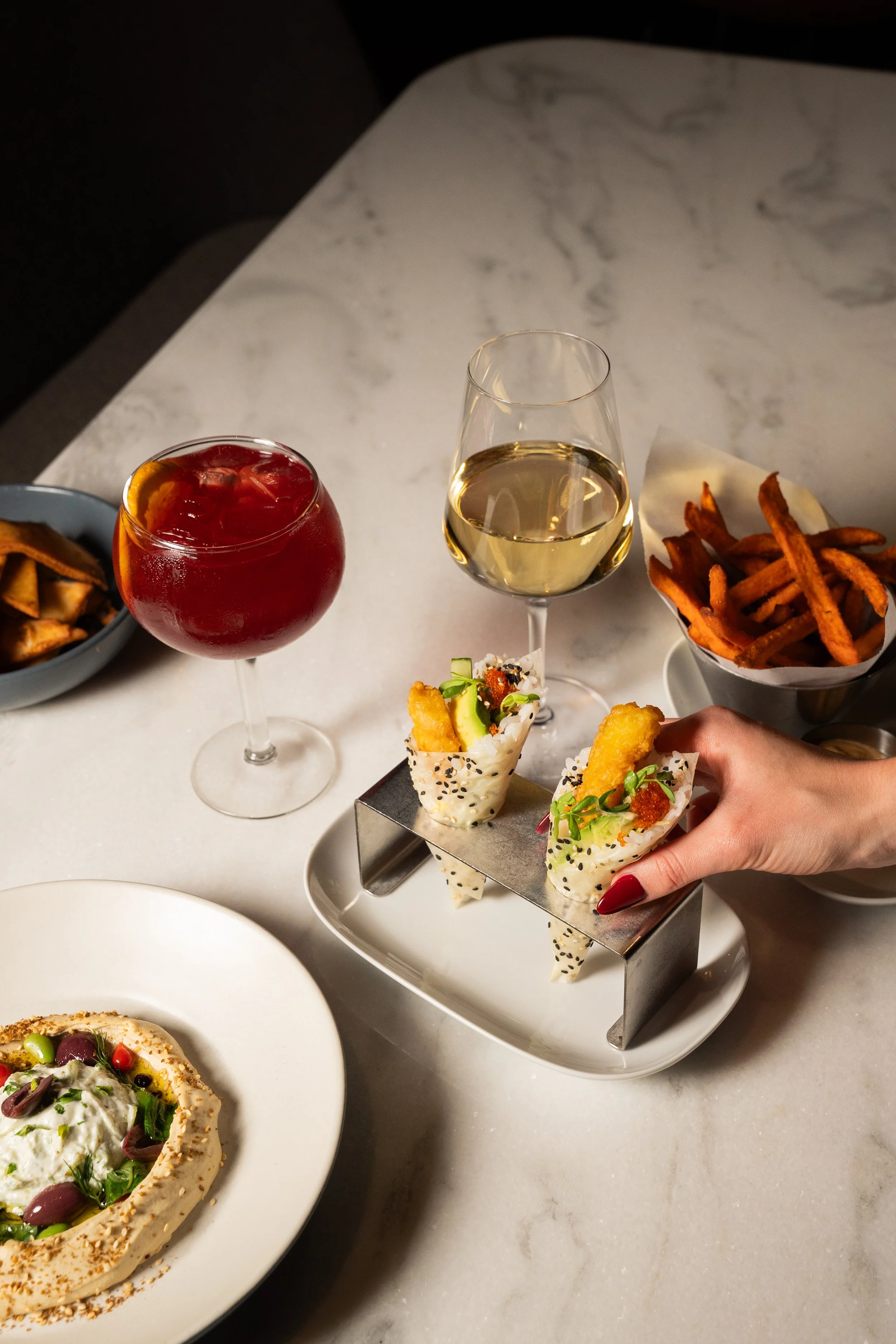 A hand holding two Asian-inspired small appetizer cones topped with garnishes, set on a white platter. Surrounding the platter are two glasses of wine, a bowl of sweet potato fries, a bowl of regular fries, and a white plate with hummus and toppings,