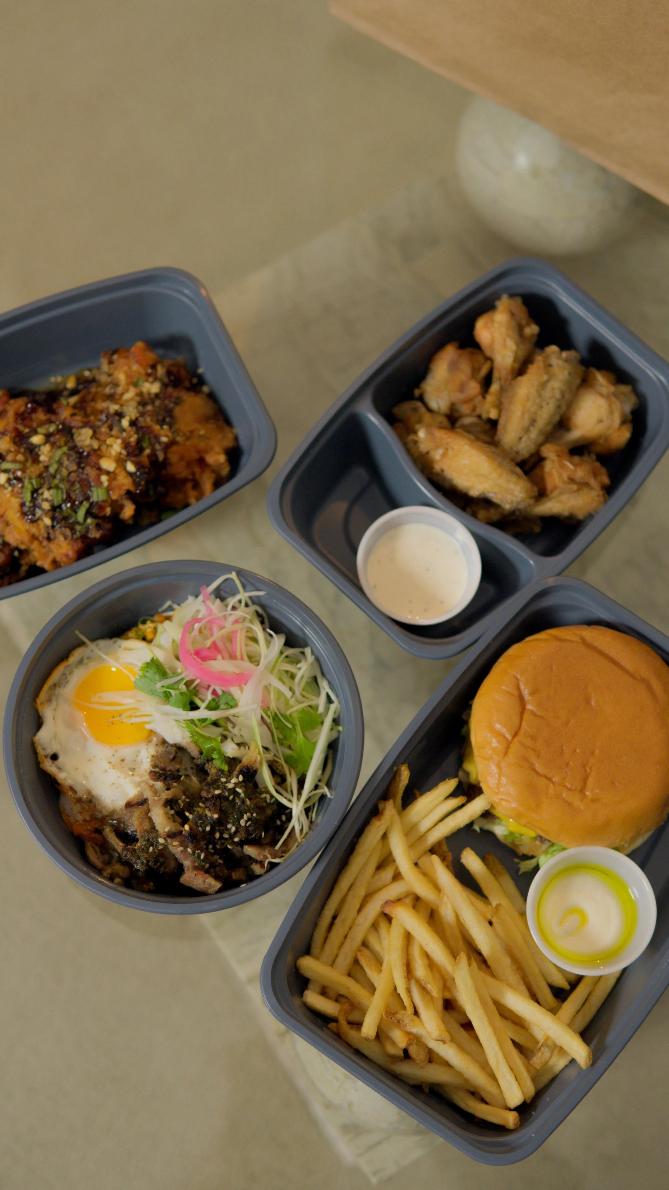 Takeout meal with a burger, fries, fried chicken wings, a dish with fried egg and salad, and a dish with baked pasta topped with crushed nuts, side sauces, all in black containers.