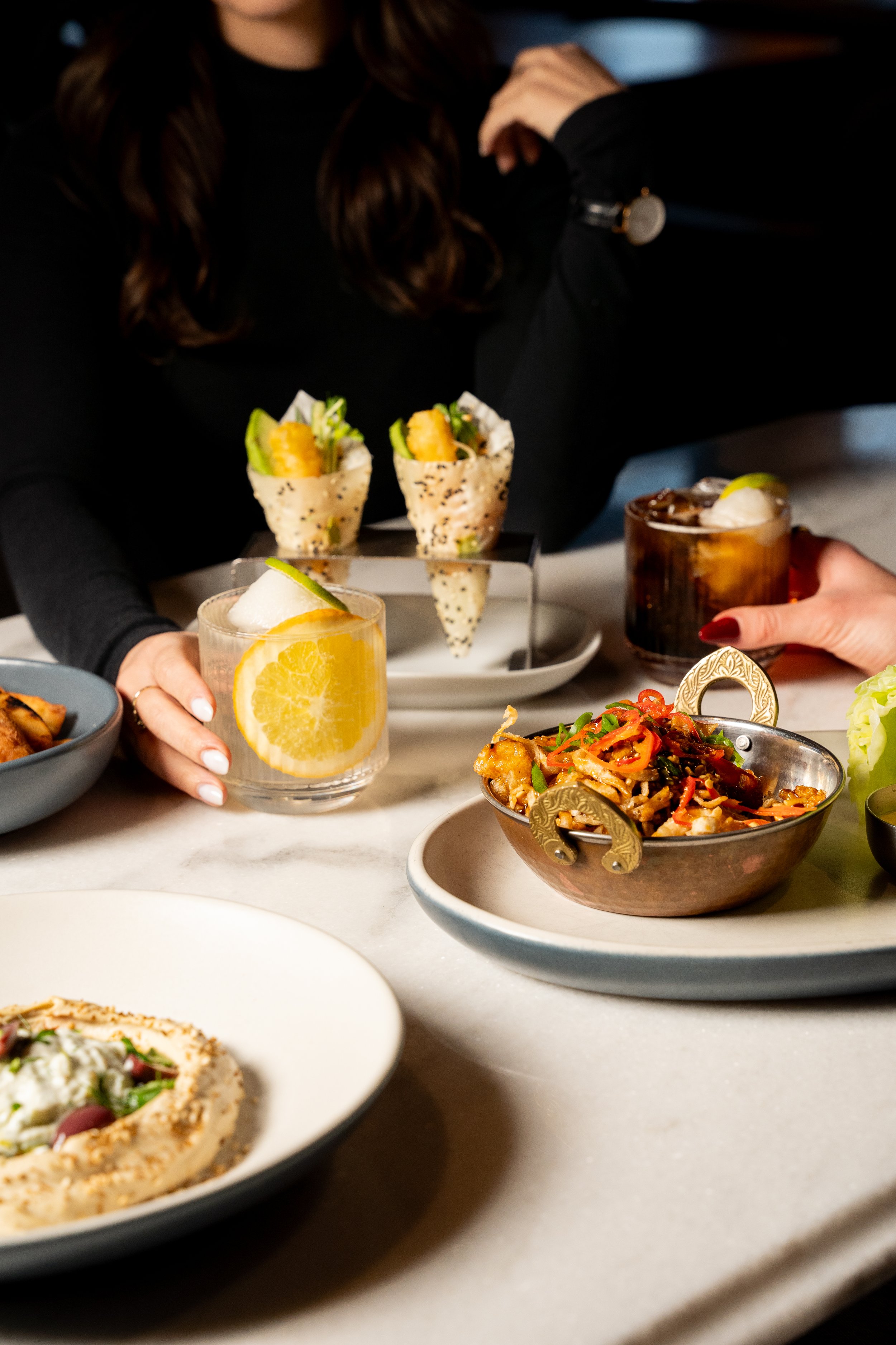 People enjoying drinks and various dishes at a restaurant table, including cocktails, a bowl of noodles, a small plate of hummus, and appetizers.