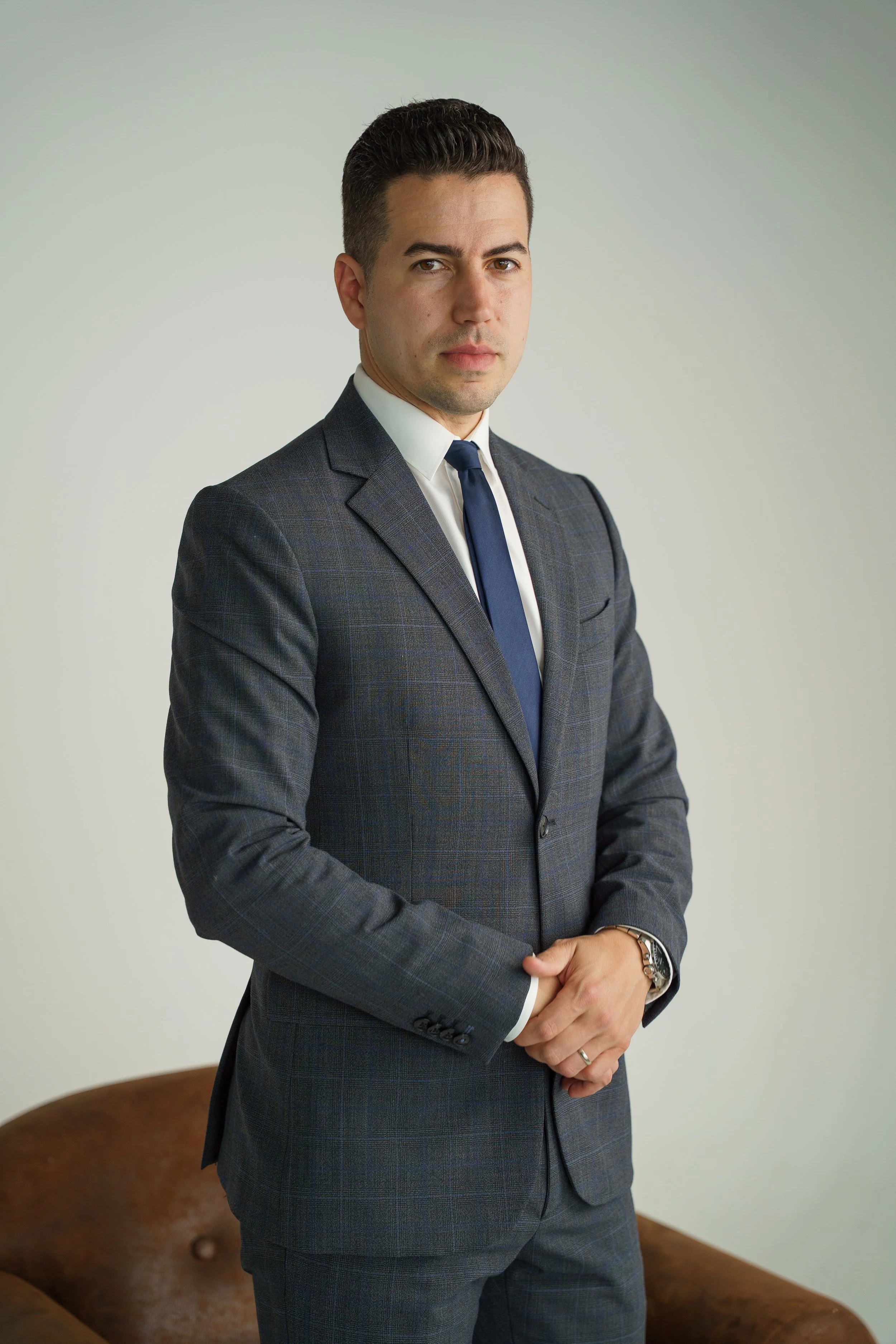 A man in a dark gray suit, white shirt, and blue tie standing with his hands clasped in front of him, looking at the camera, against a plain light-colored background.