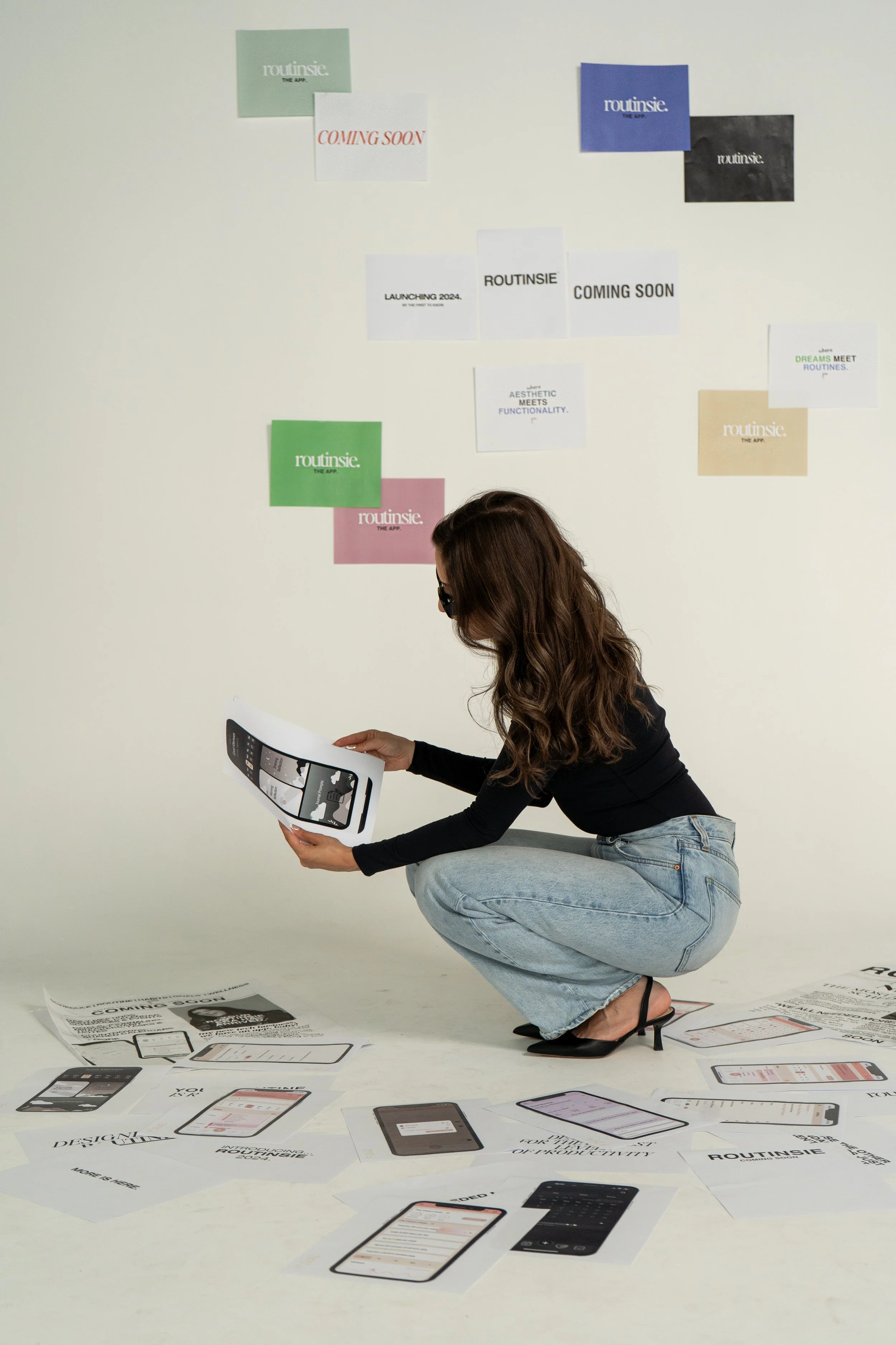 A woman with long brown hair, wearing glasses, a black top, light blue jeans, and black heels, crouching on the floor while holding and examining a paper with phone app designs. Multiple sheets of paper with app interfaces are scattered on the floor 