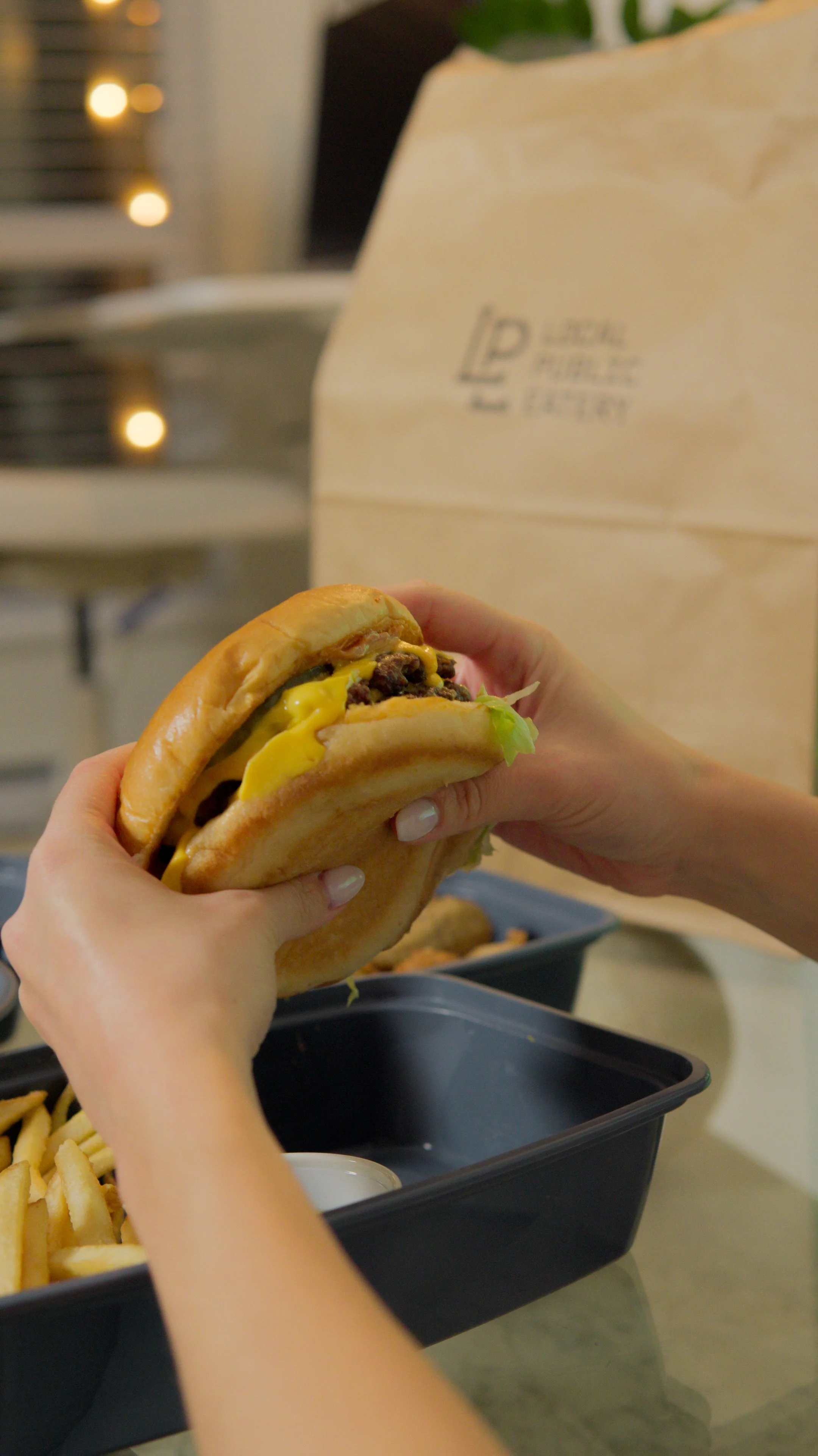 Person holding a cheeseburger with fries and a drink in the background.