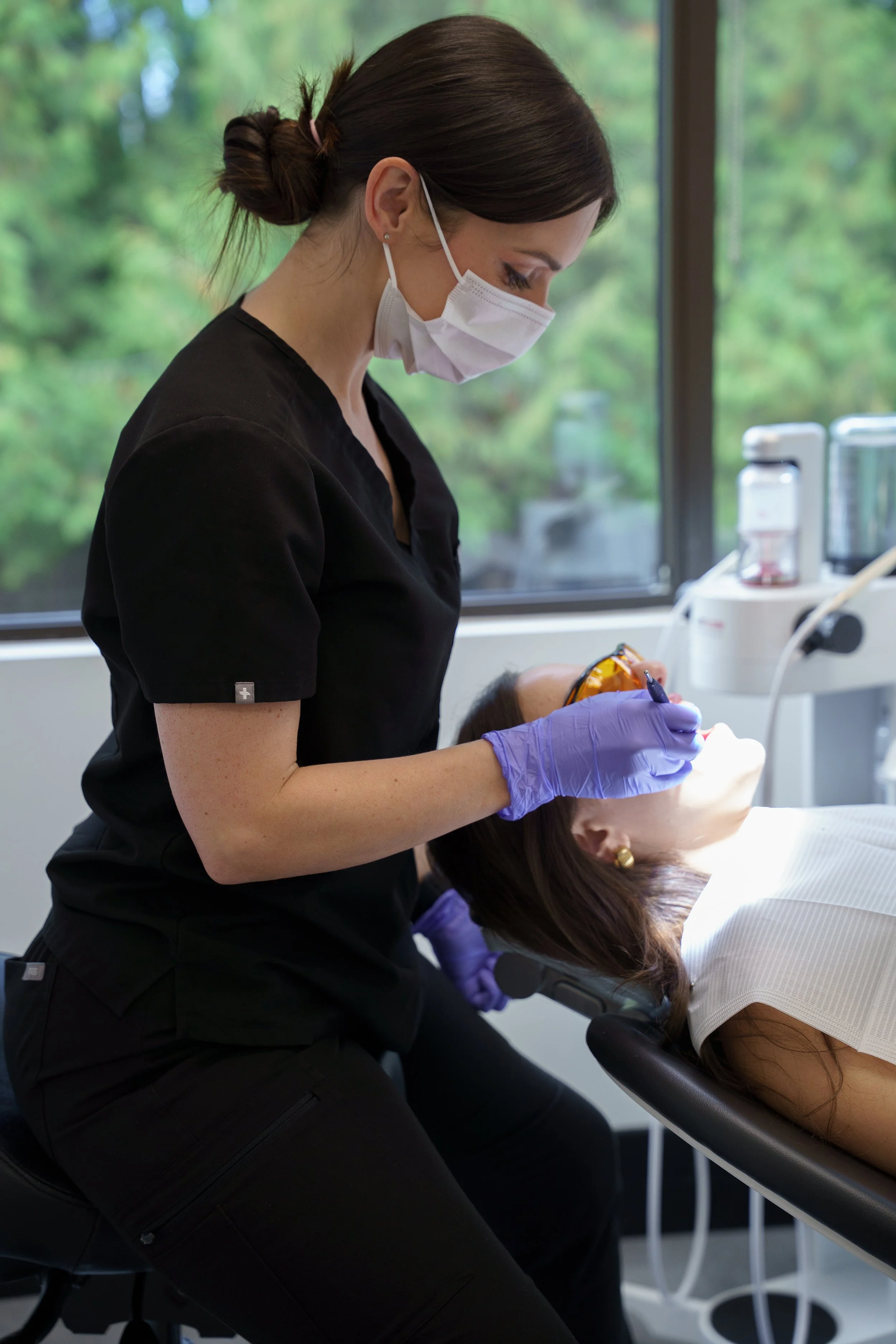 Dental professional in black scrubs and purple gloves examines a patient lying in a dental chair with a mask and sunglasses, next to dental equipment and a window with a view of green trees.