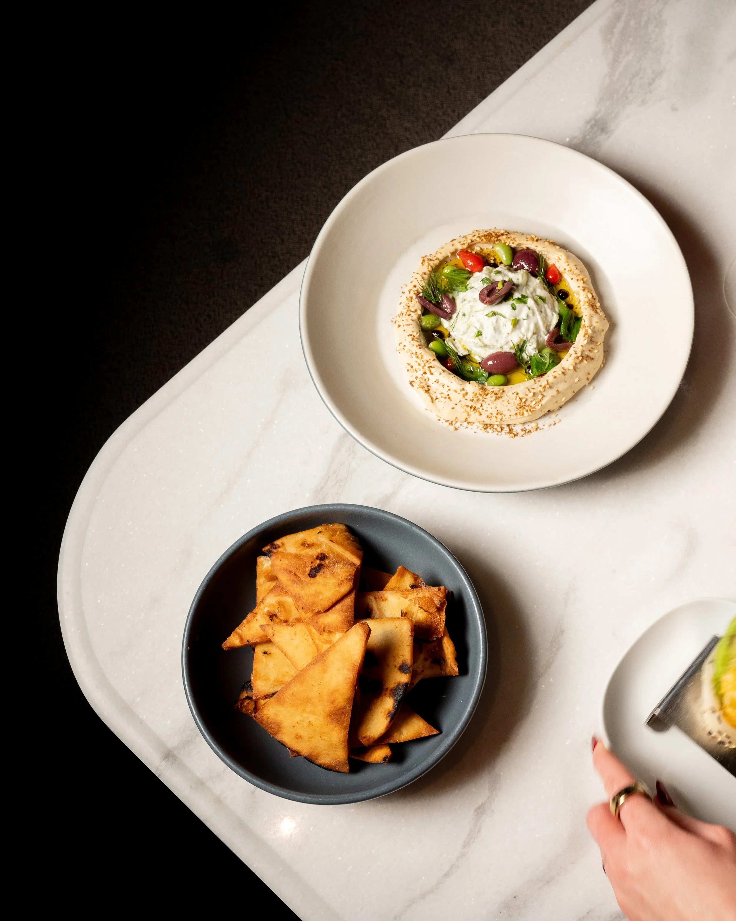 A white plate with a creamy dip or spread garnished with herbs, olives, and cherry tomatoes, surrounded by a ring of toasted bread or crackers. Next to it, a black bowl contains triangular pieces of grilled pita or flatbread. A person's hand with a r
