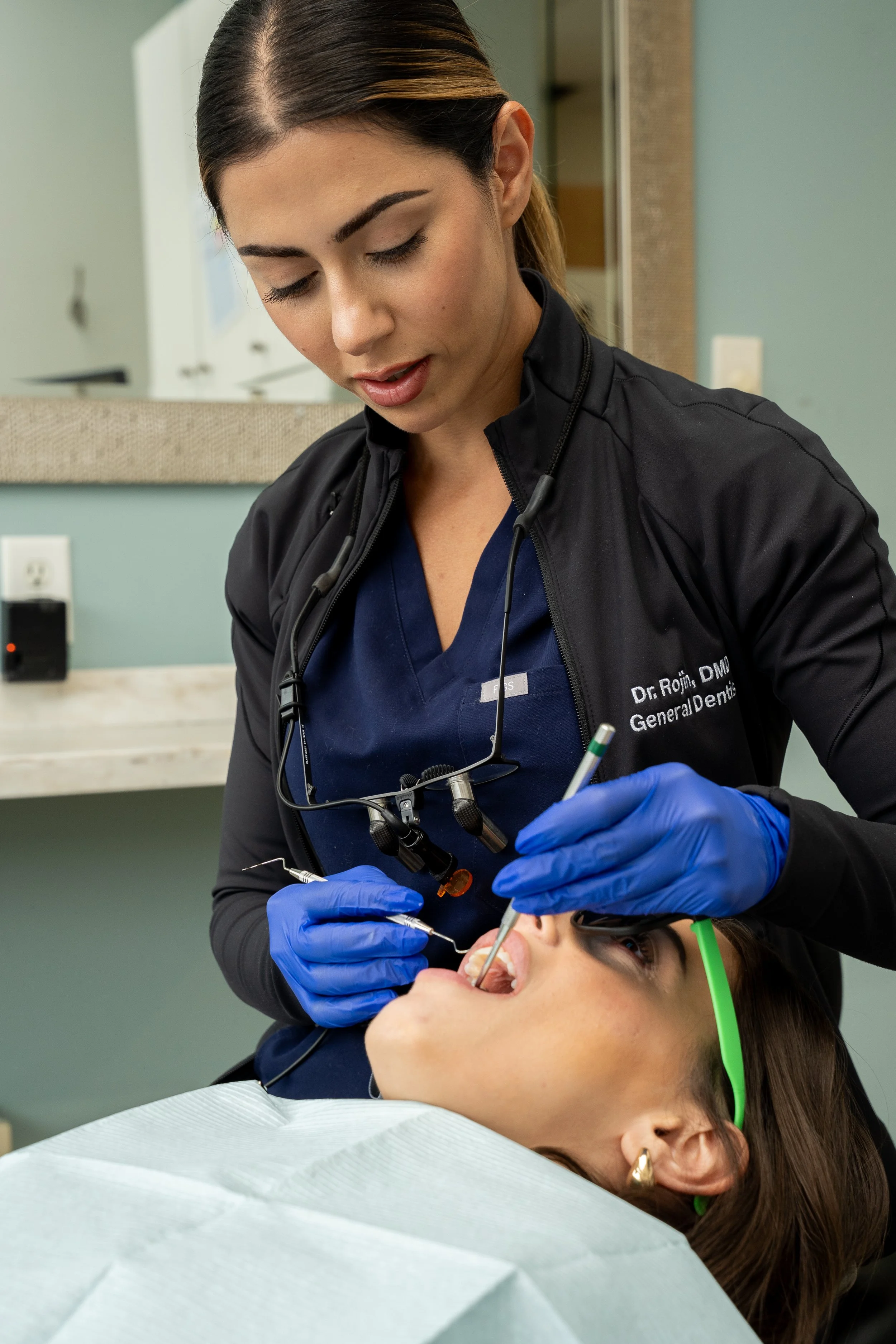 A female dentist with dark hair, wearing blue gloves, a dark jacket, and a blue uniform, performs a dental procedure on a patient lying in a dental chair.