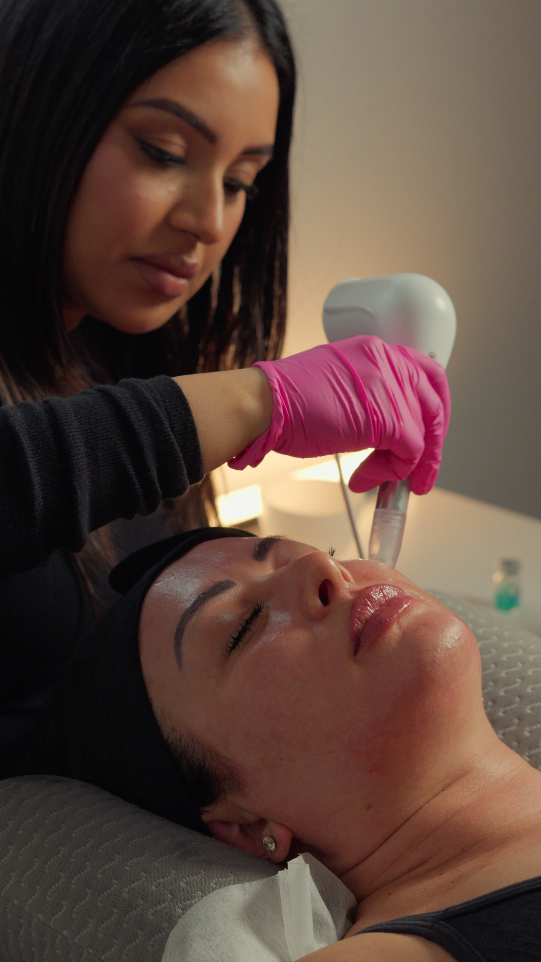A woman is receiving a cosmetic or dermatological treatment on her face from a professional wearing pink gloves.