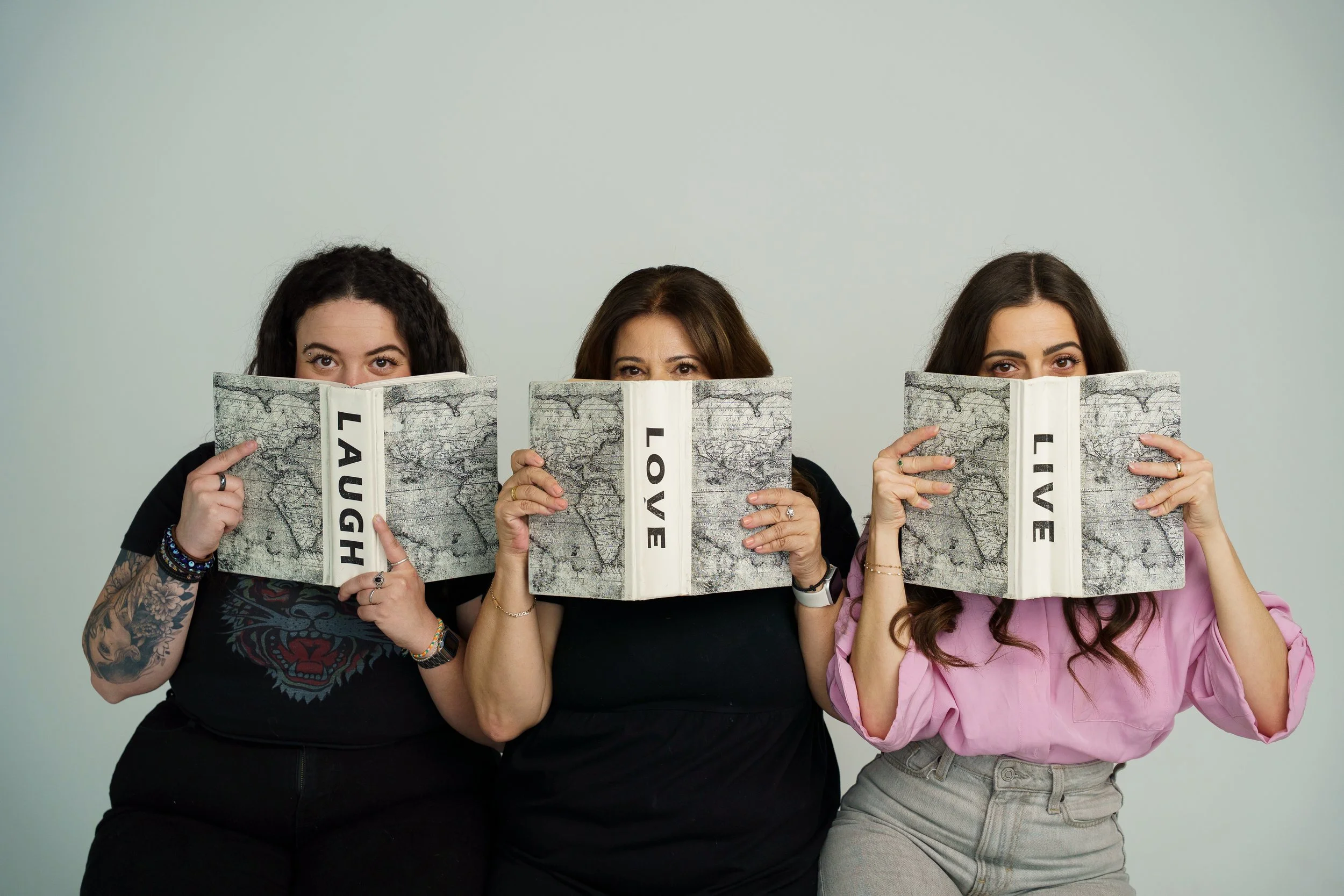Three women holding open books with the words 'LAUGH,' 'LOVE,' and 'LIVE' printed on the spines, partially covering their faces, against a plain background.