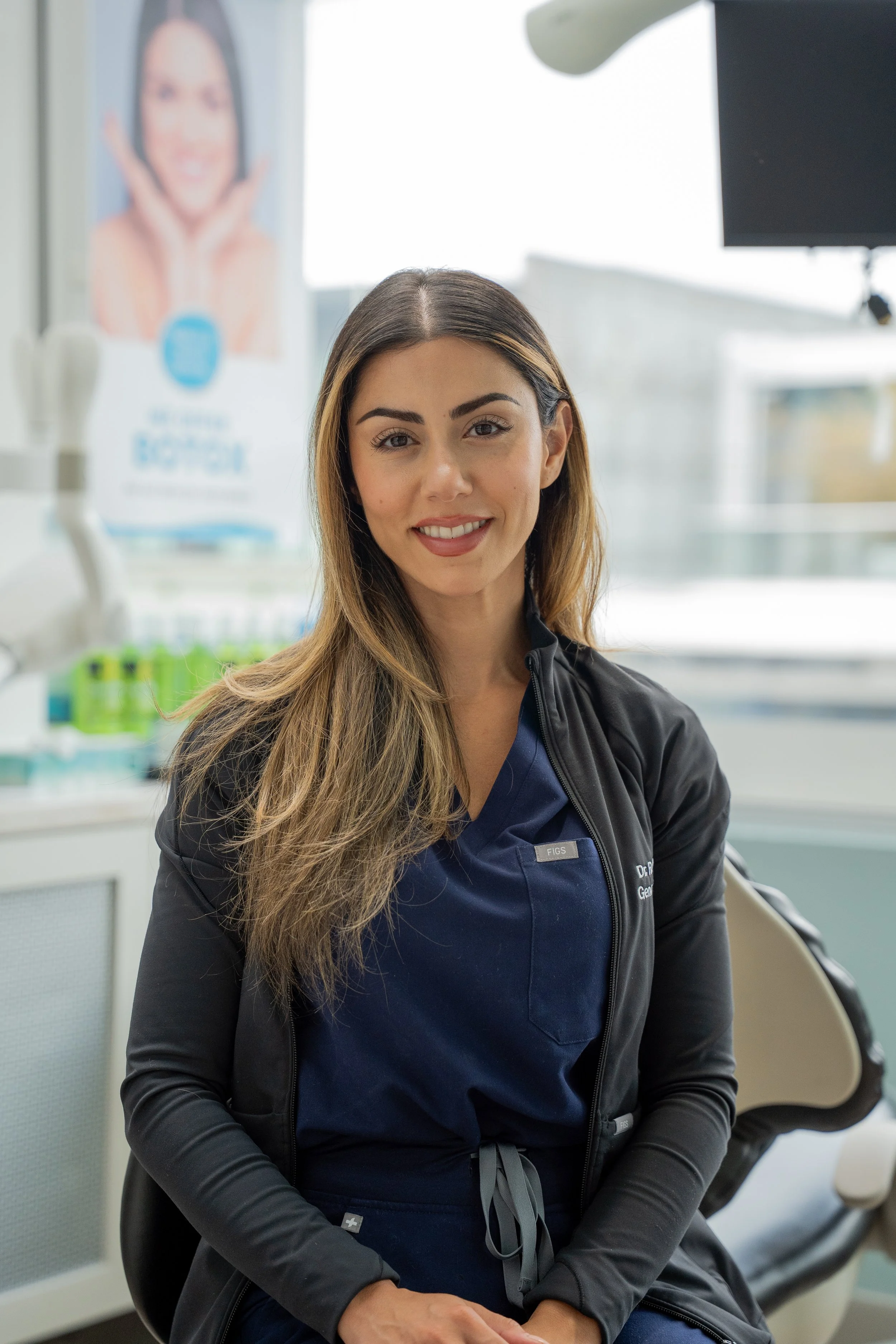 A smiling woman with long brown hair and a black jacket seated in a dental or medical office.
