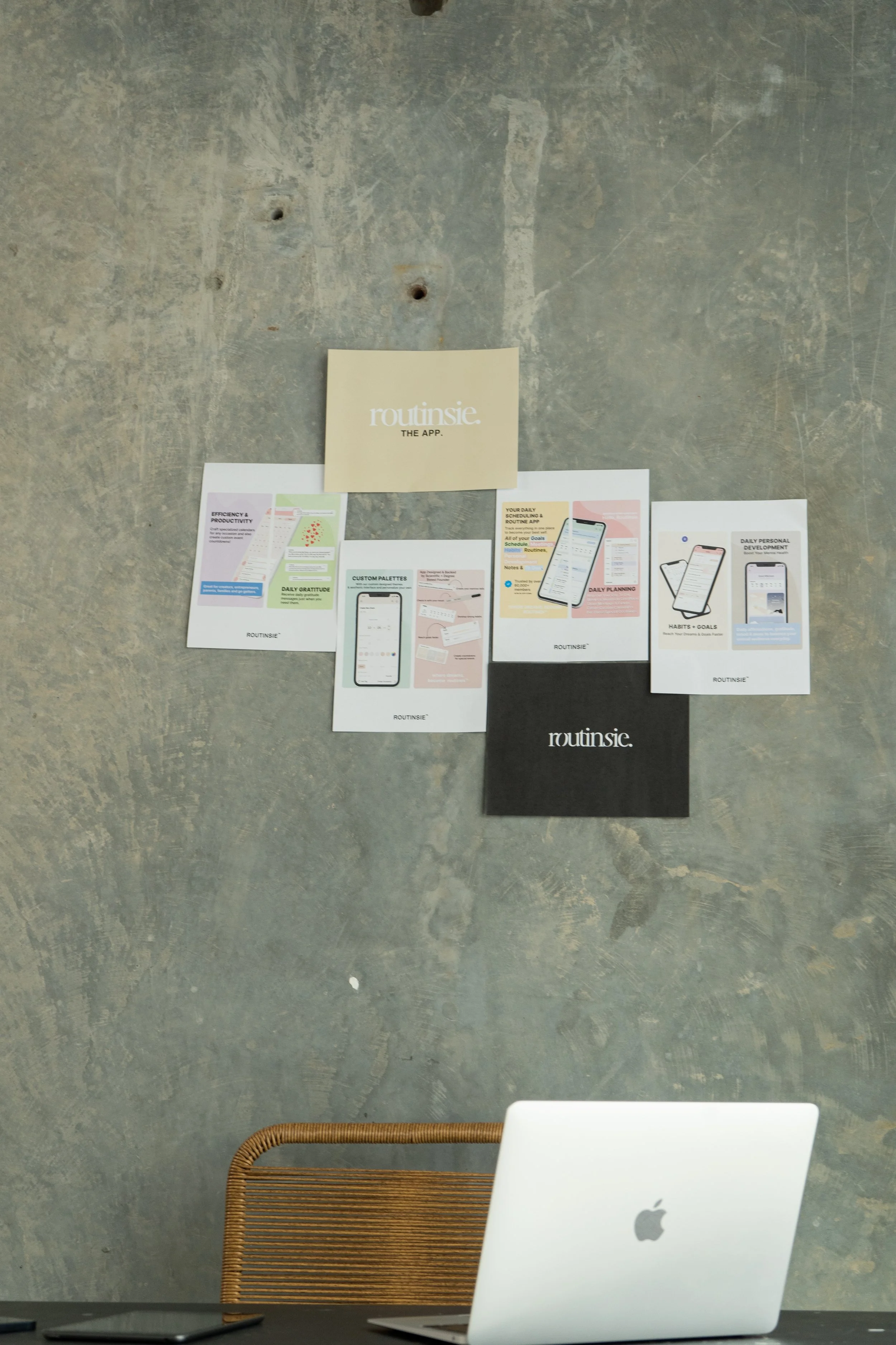 Desk with a silver Apple MacBook and a black smartphone in front of a concrete wall with various printed marketing materials for Routines, an app, pinned to the wall.