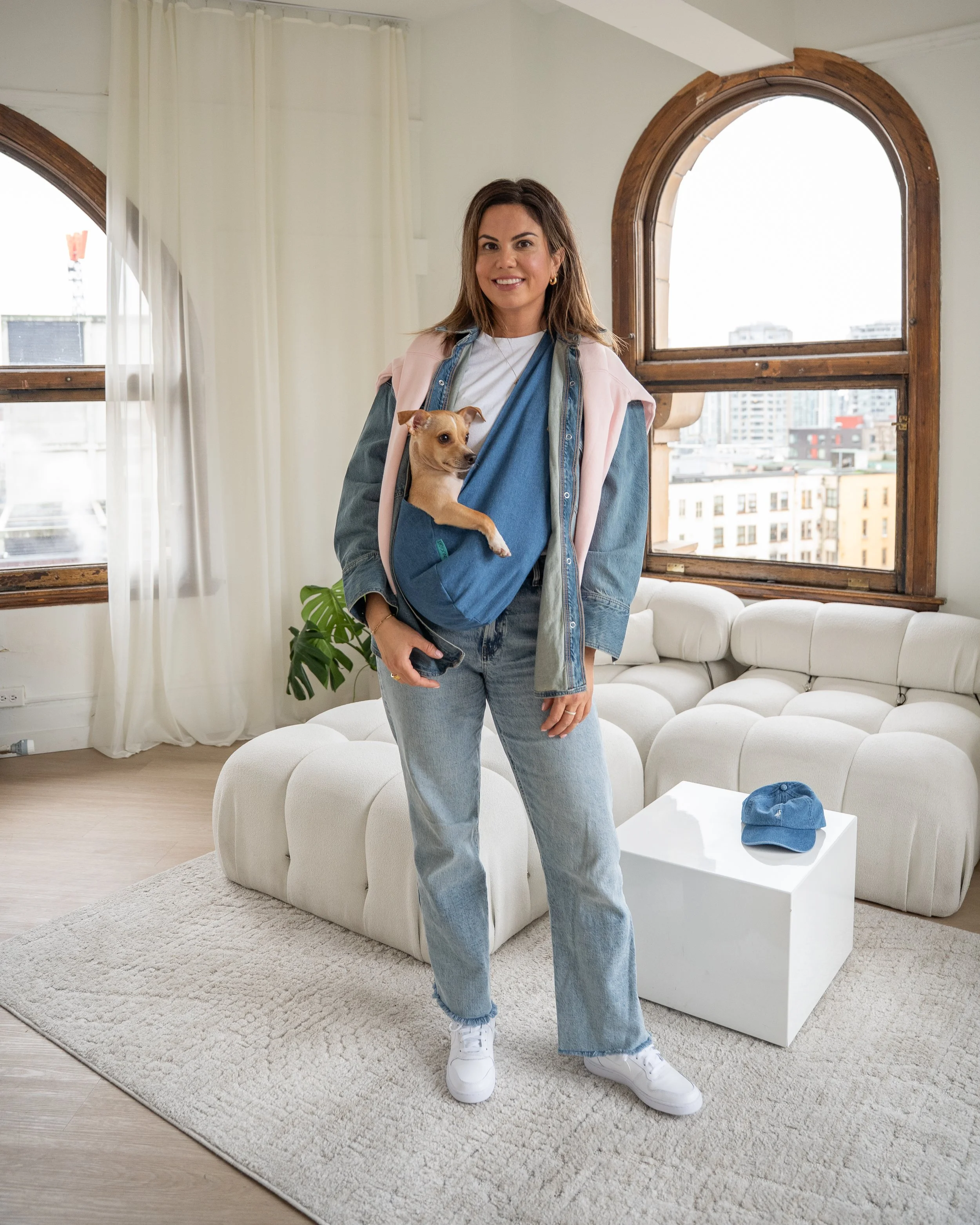 Woman wearing casual denim and white sneakers standing in a modern living room with large windows, holding a small dog in a blue dog carrier.