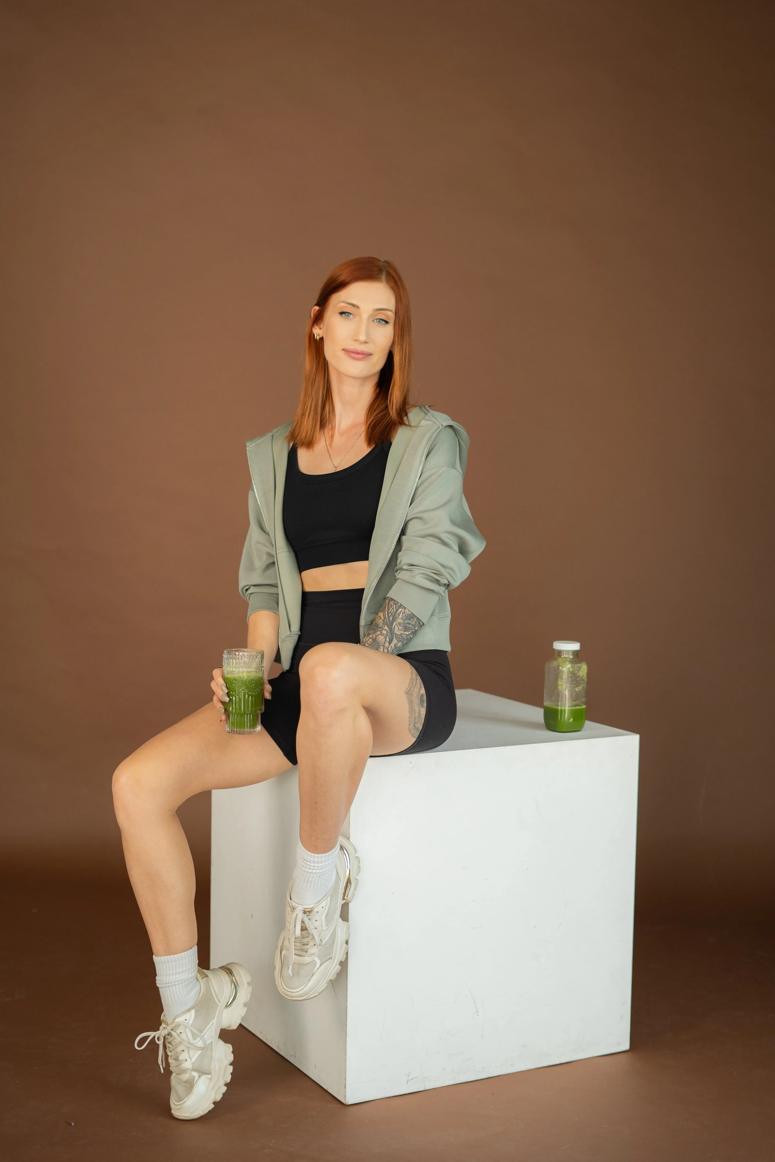 A woman with red hair and tattoos sitting on a white cube, holding a glass of green juice, with a bottle of green juice nearby, against a brown background.