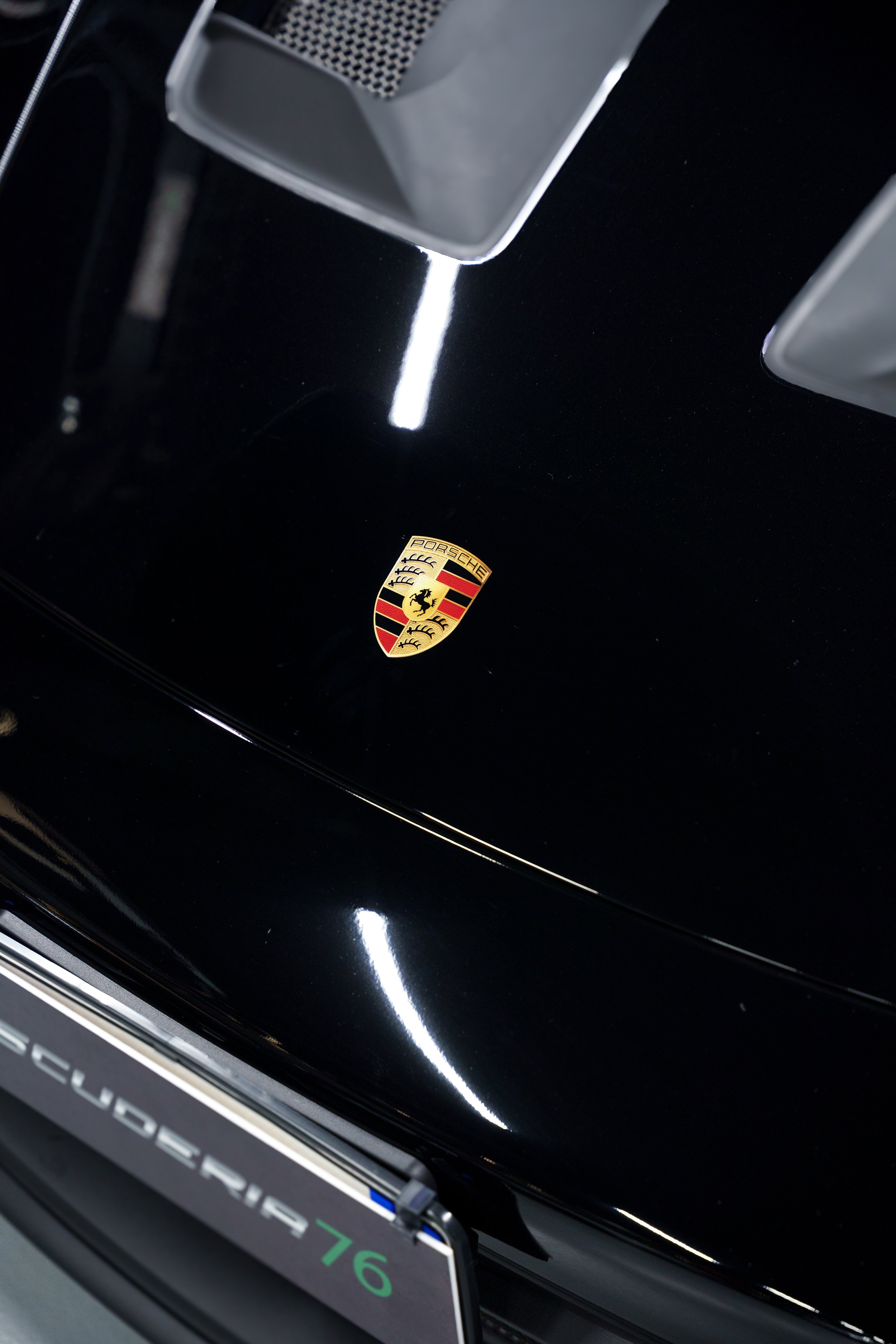 Close-up of a black Porsche car hood showing the Porsche emblem and part of the license plate with the number 76.