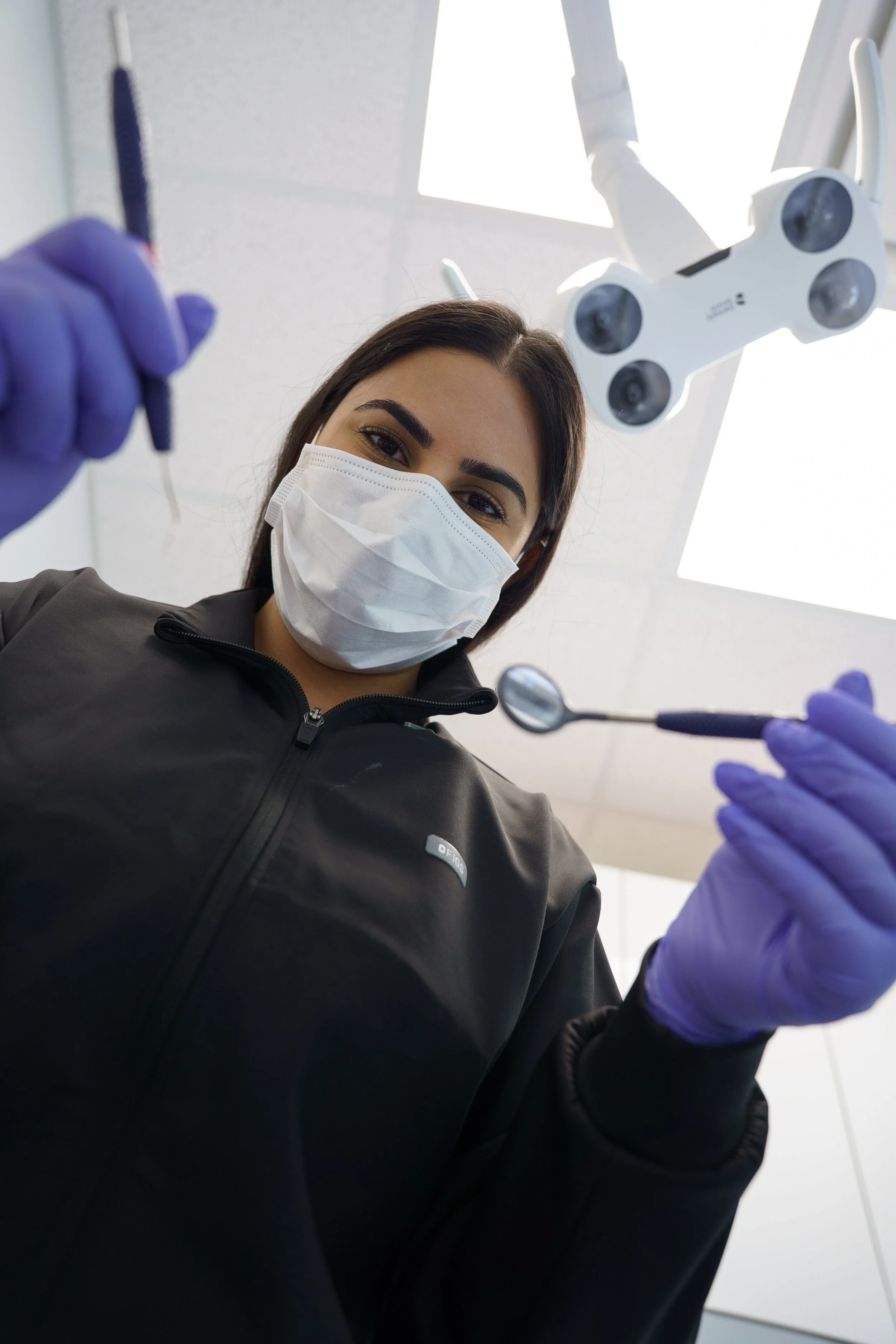 Dental professional wearing a face mask and gloves, looking down at dental tools, with dental light and ceiling in background.