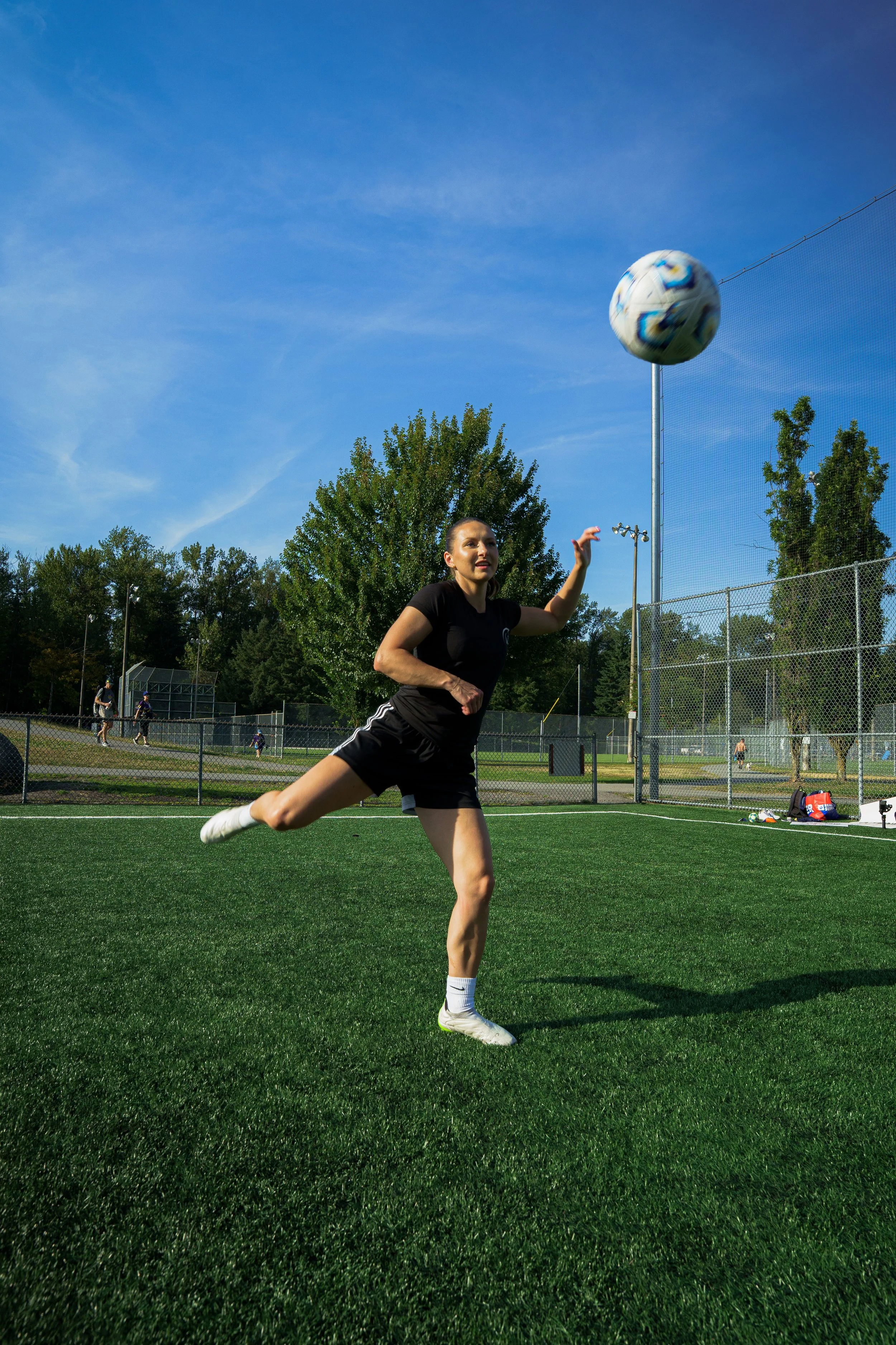 A woman playing soccer on a green field, about to kick a soccer ball into the air. She is wearing a black sports outfit and white shoes, with a blue sky and trees in the background.