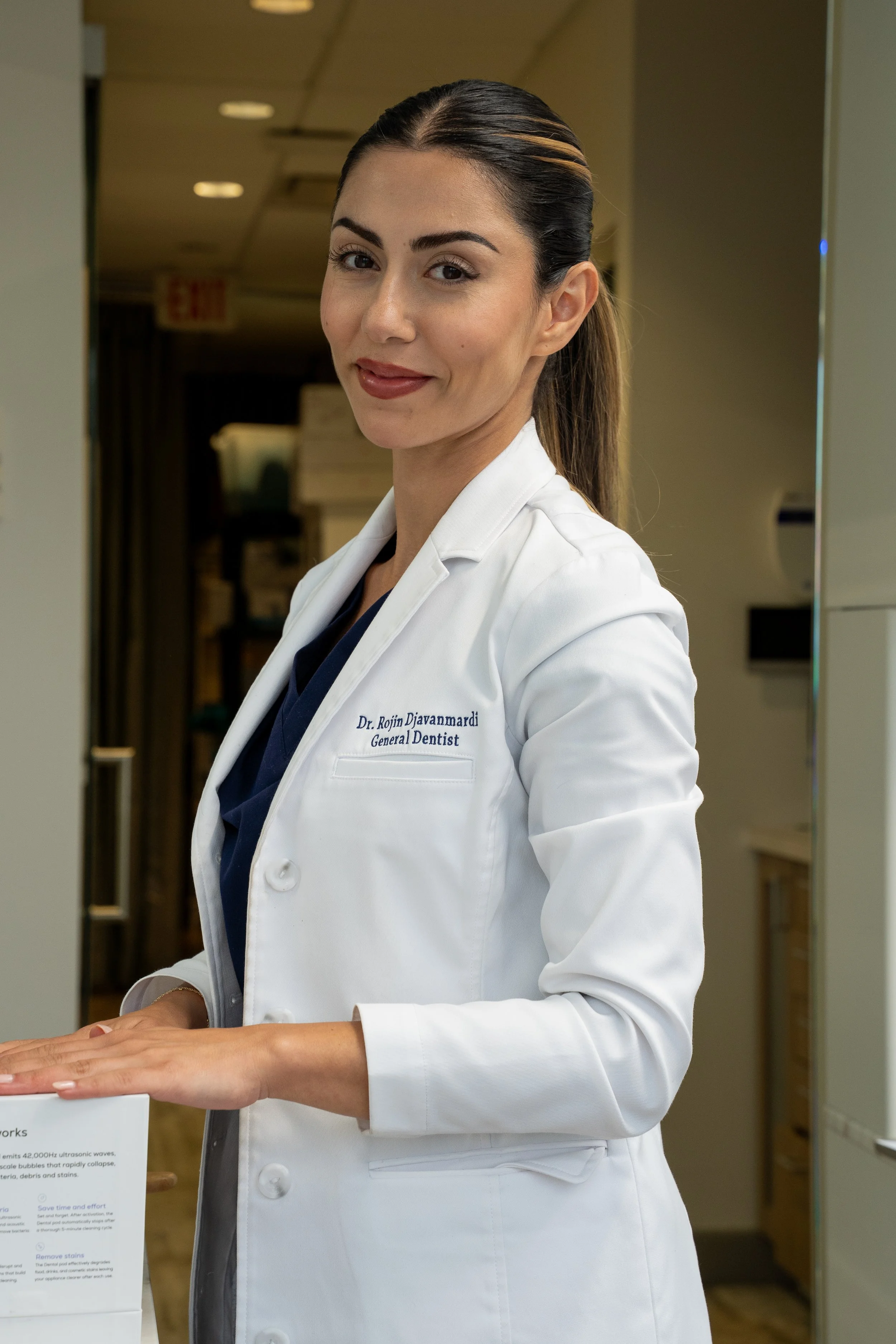 A woman wearing a white coat with 'Dr. Rojin Djanvardi, General Dentist' embroidered on it, standing in a professional setting, smiling at the camera.
