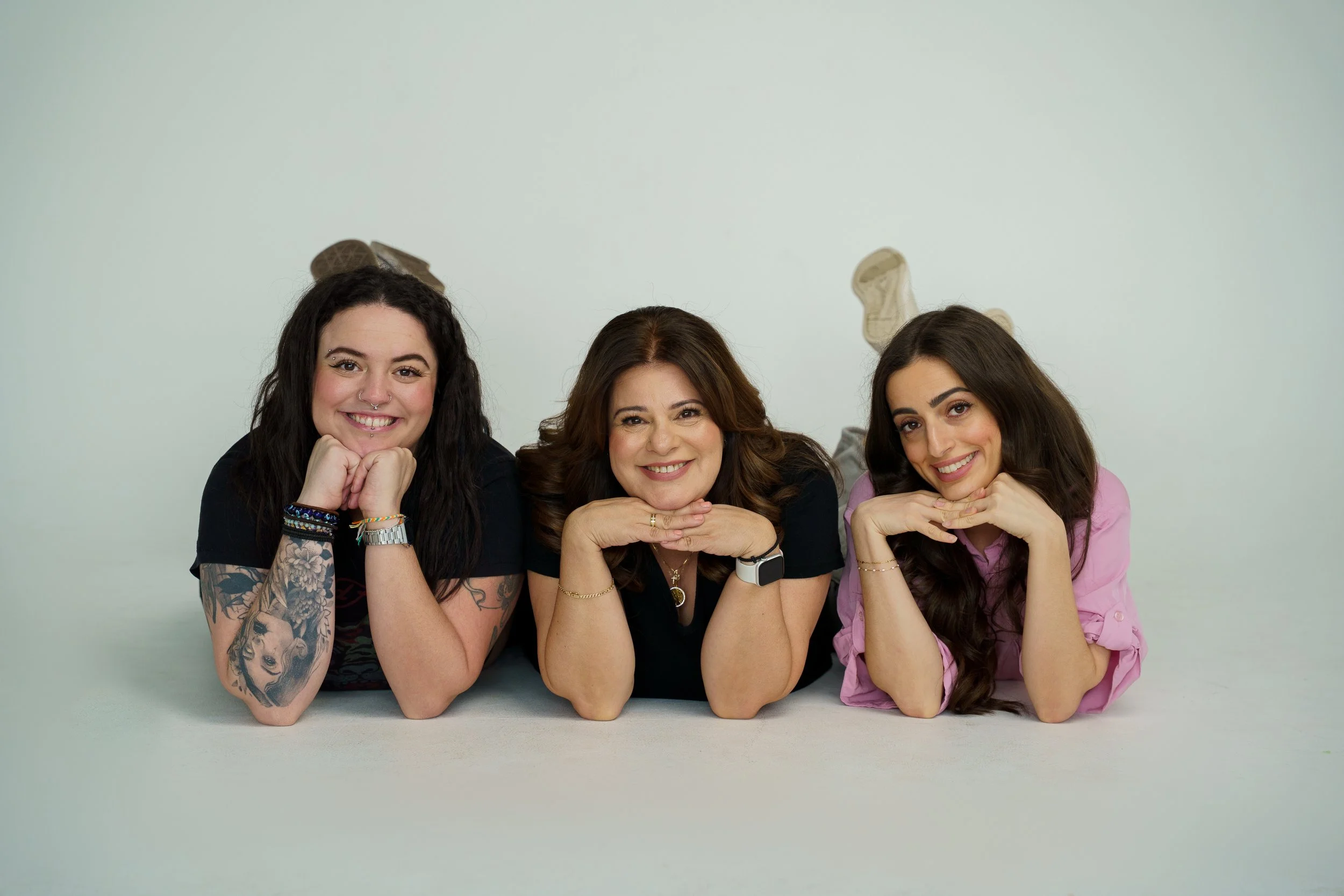 Three women lying on the floor with their heads resting on their hands, smiling at the camera, against a plain light background.