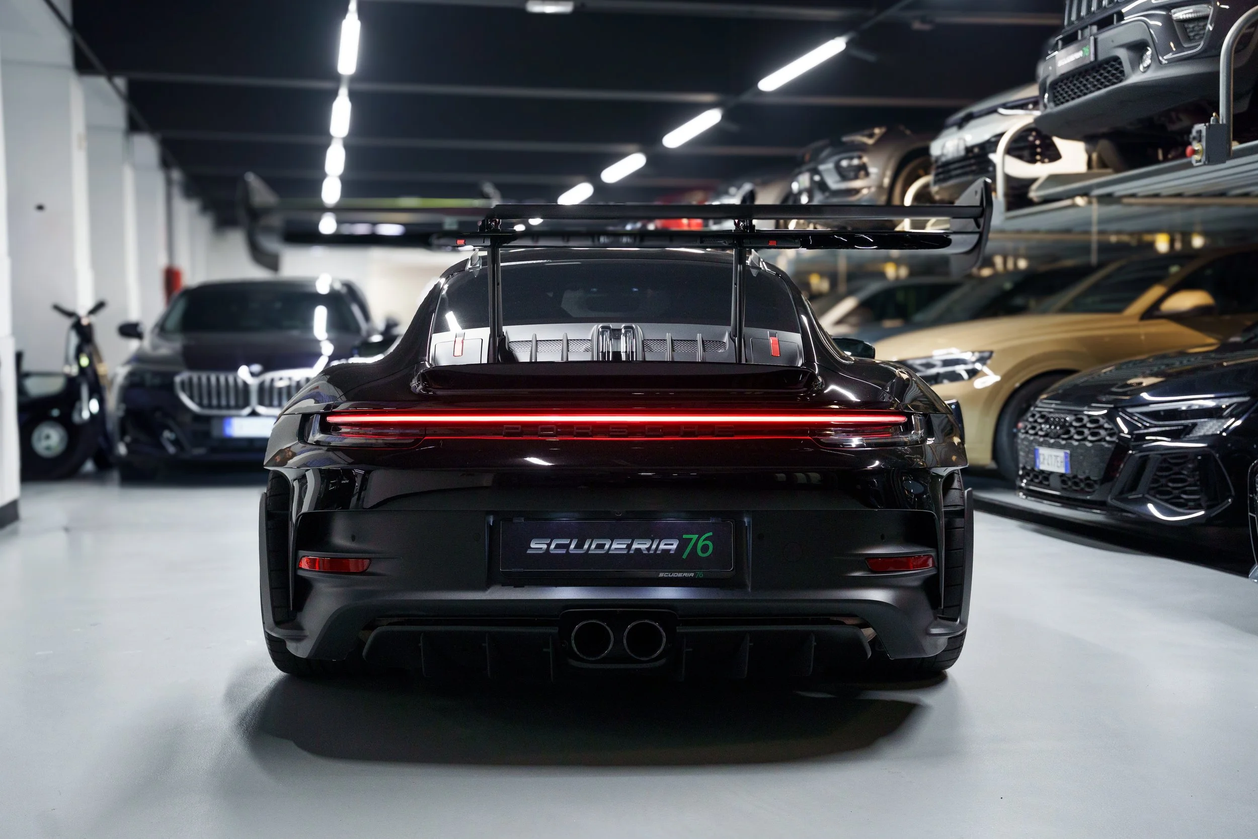 Black Porsche 911 with a large rear wing inside a multi-level car garage, surrounded by other luxury cars and SUVs.